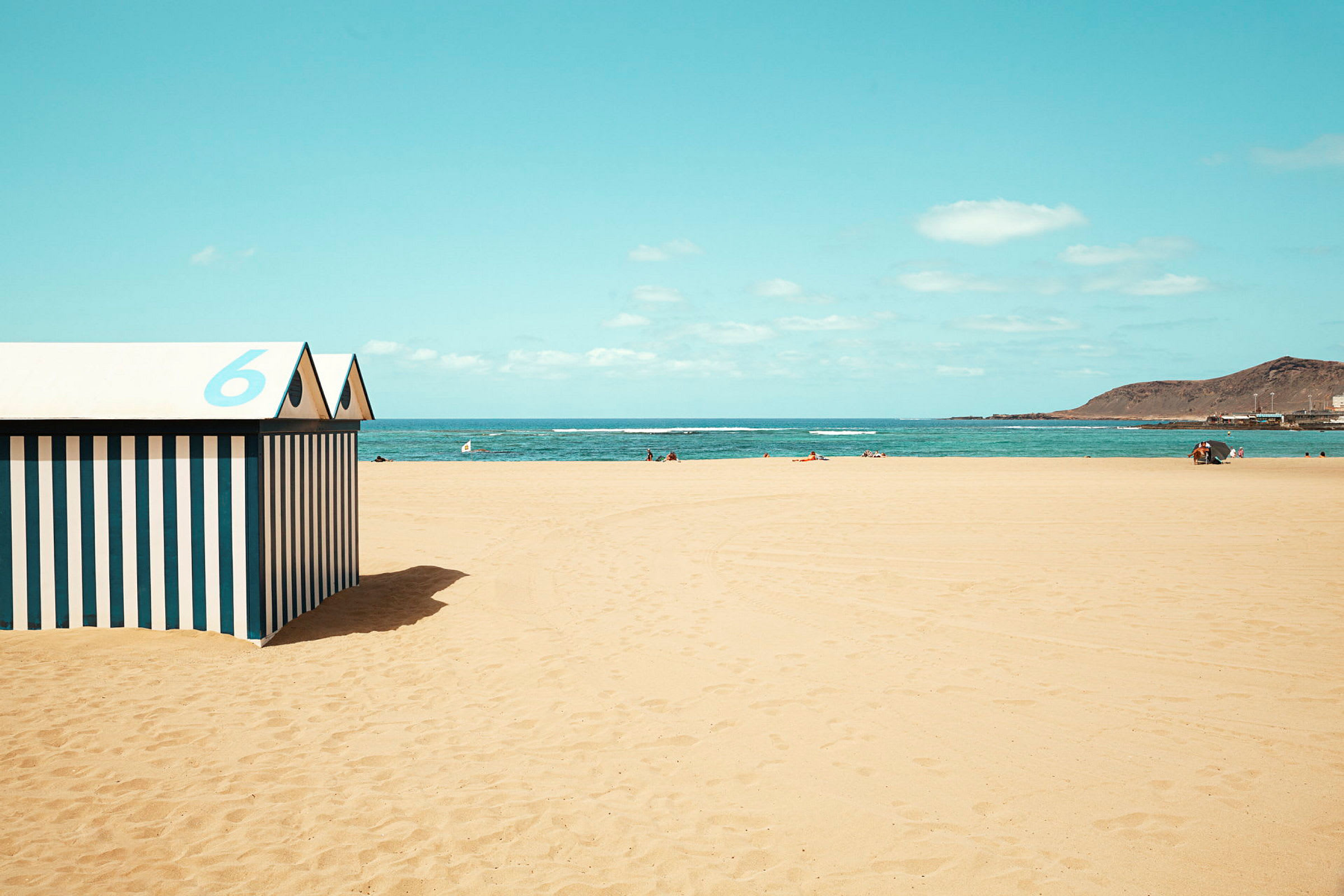 Strand med gul sand, blått hav og himmel, samt et stripete strandhus med nummer 6 i forgrunnen. (AI-generert tekst)