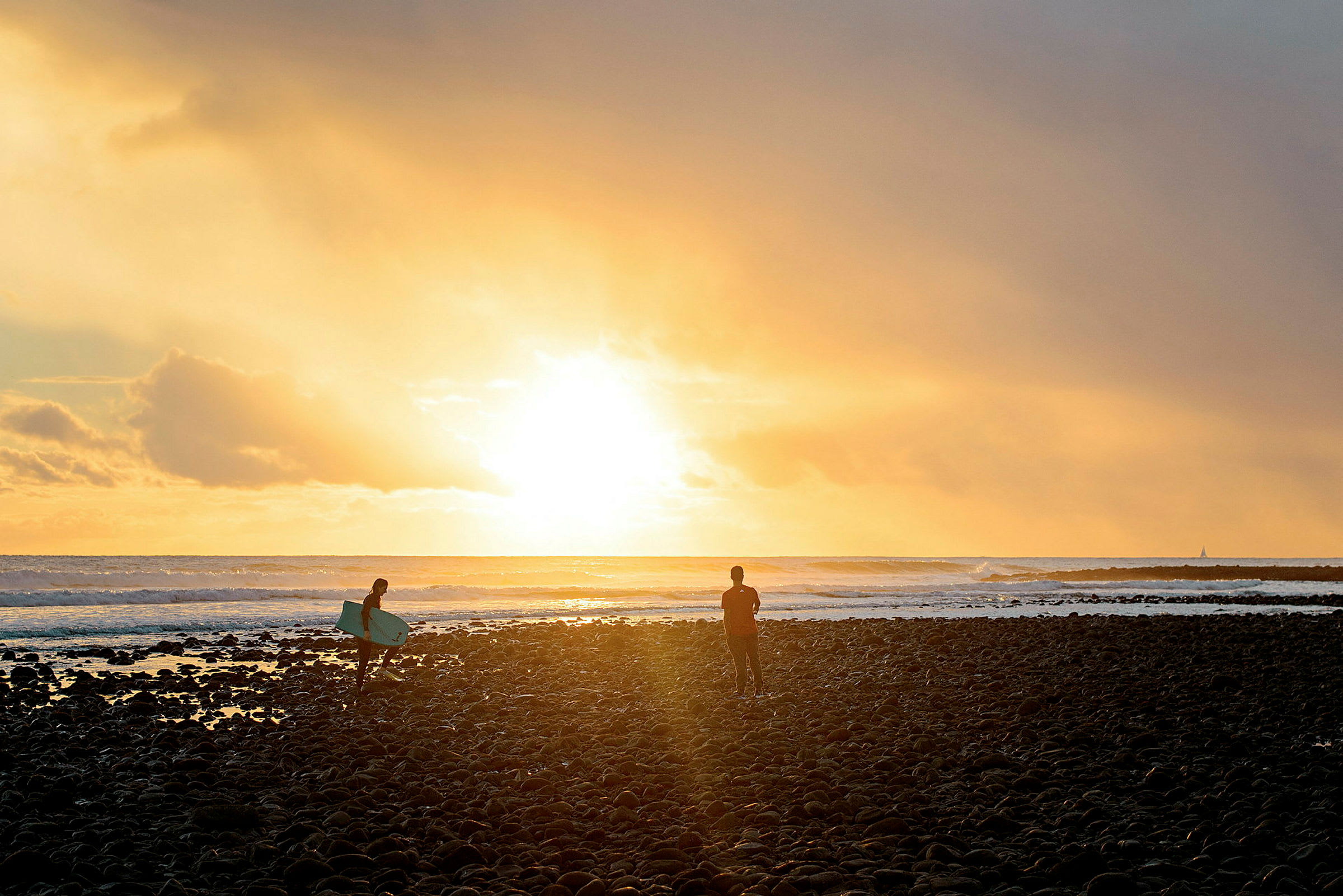 En solnedgang over havet med to personer på en steinete strand, den ene med et surfebrett. (AI-generert tekst)