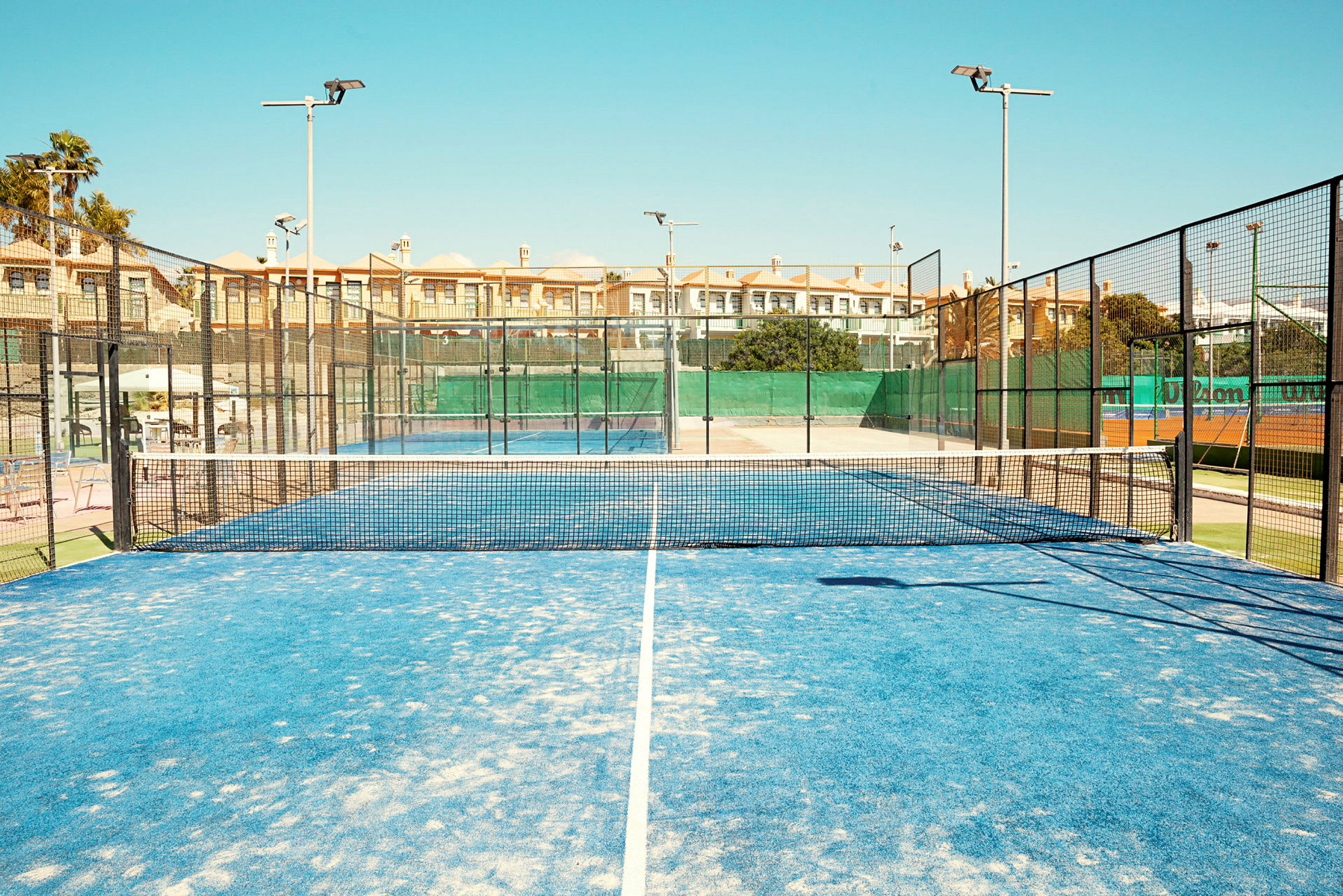 En padelbane med blått underlag og nett i midten, omgitt av gjerder og bygninger i bakgrunnen. (AI-generert tekst)