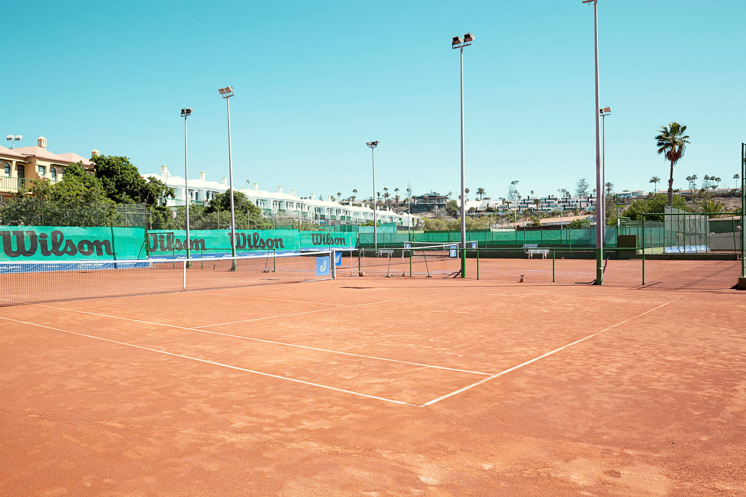 Tennisbaner med grusunderlag, omgitt av nett og lysmaster. (AI-generert tekst)