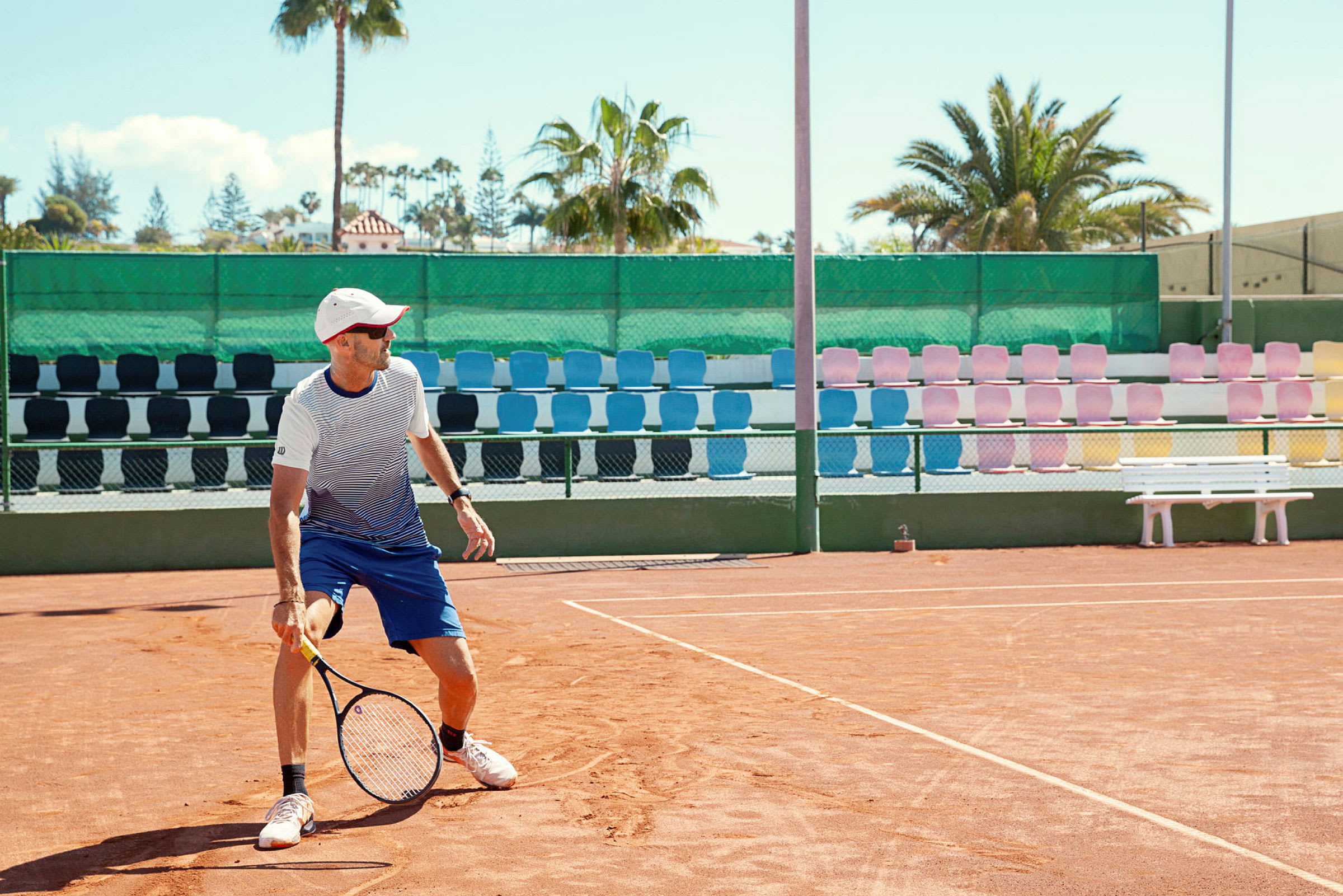 En tennisbane med en spillere som holder en racket, omgitt av fargerike seter og palmer i bakgrunnen. (AI-generert tekst)