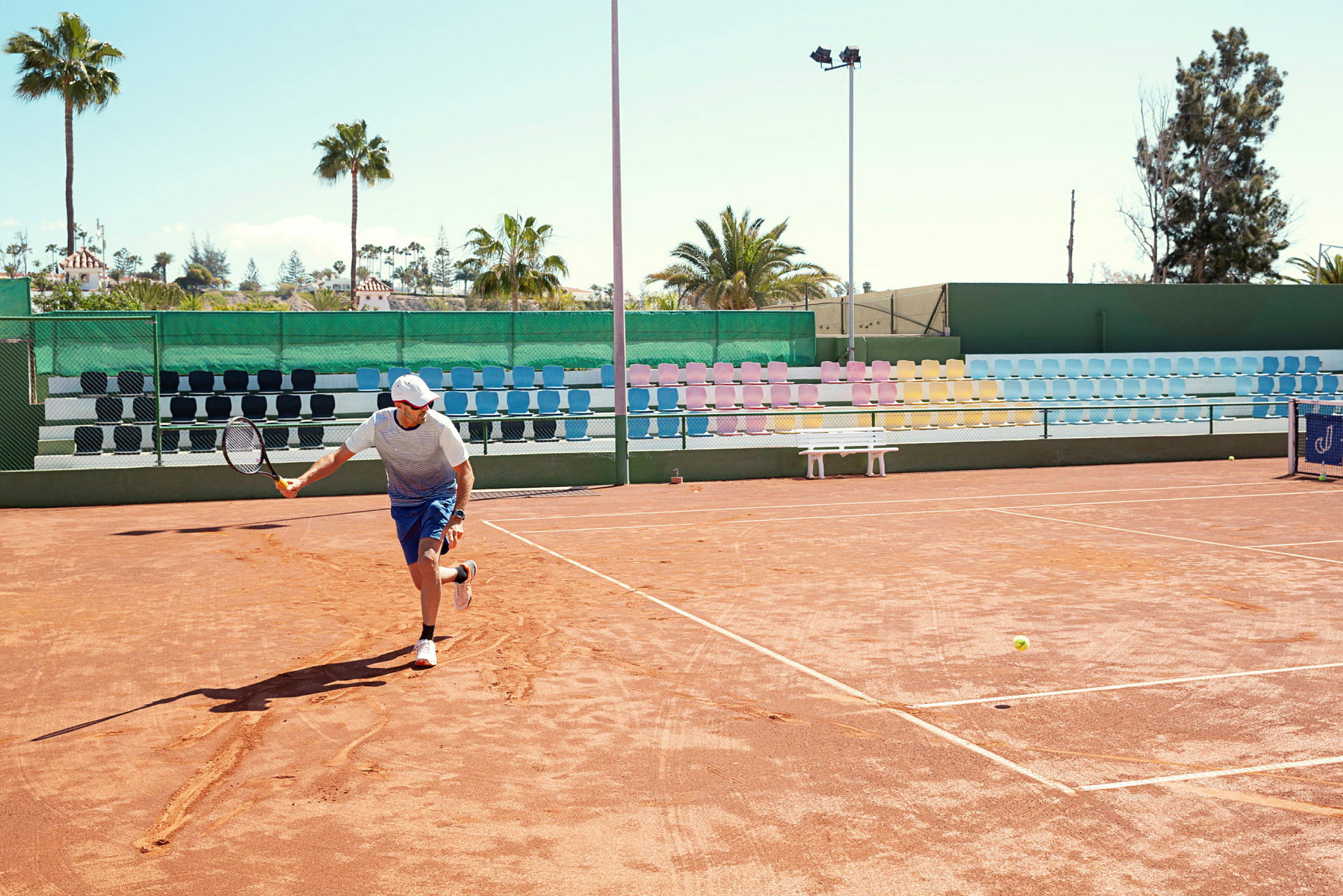 En person spiller tennis på en grusbane med fargerike tribuneseter og palmer i bakgrunnen. (AI-generert tekst)