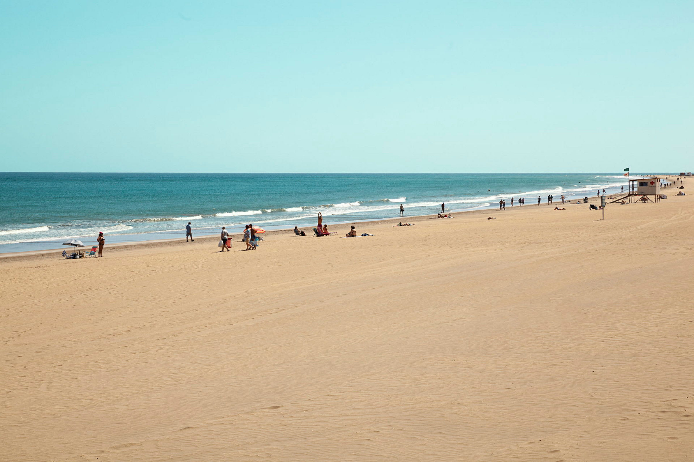 En lang sandstrand med mennesker som nyter solen og havet, samt en livredningsstasjon. (AI-generert tekst)