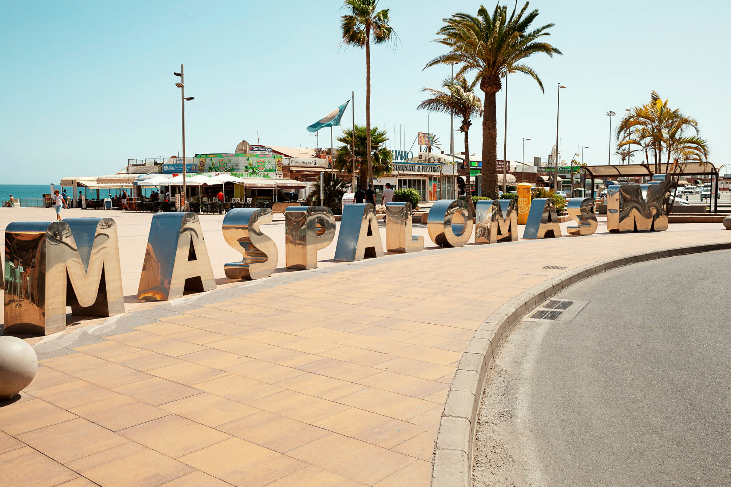 Store, blanke bokstaver danner ordet "MASPALOMAS" på en fortau ved et torg med palmer. (AI-generert tekst)