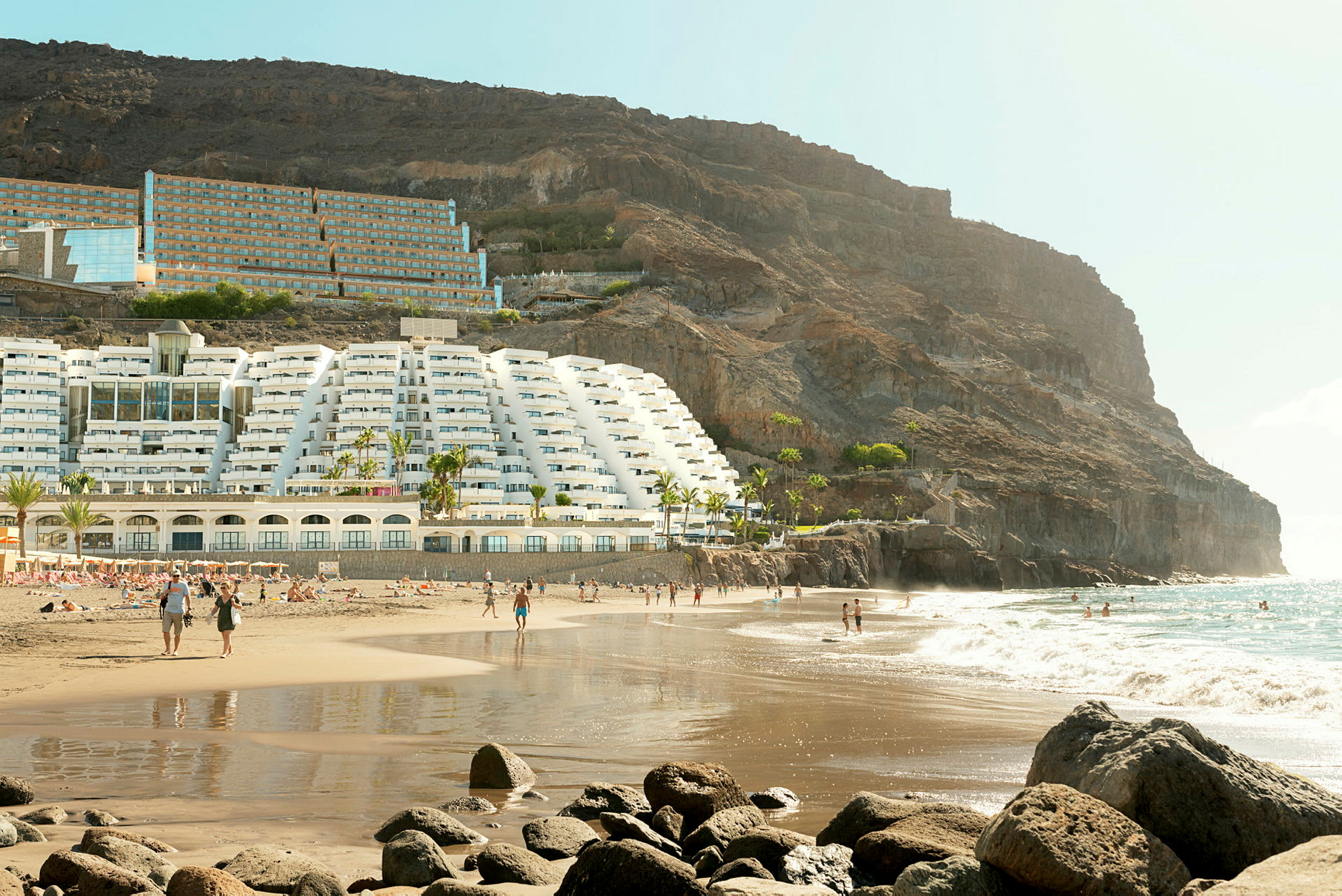 Strand med hotellbygning ved en skråning og folk som beveger seg langs sanden og vannet. (AI-generert tekst)