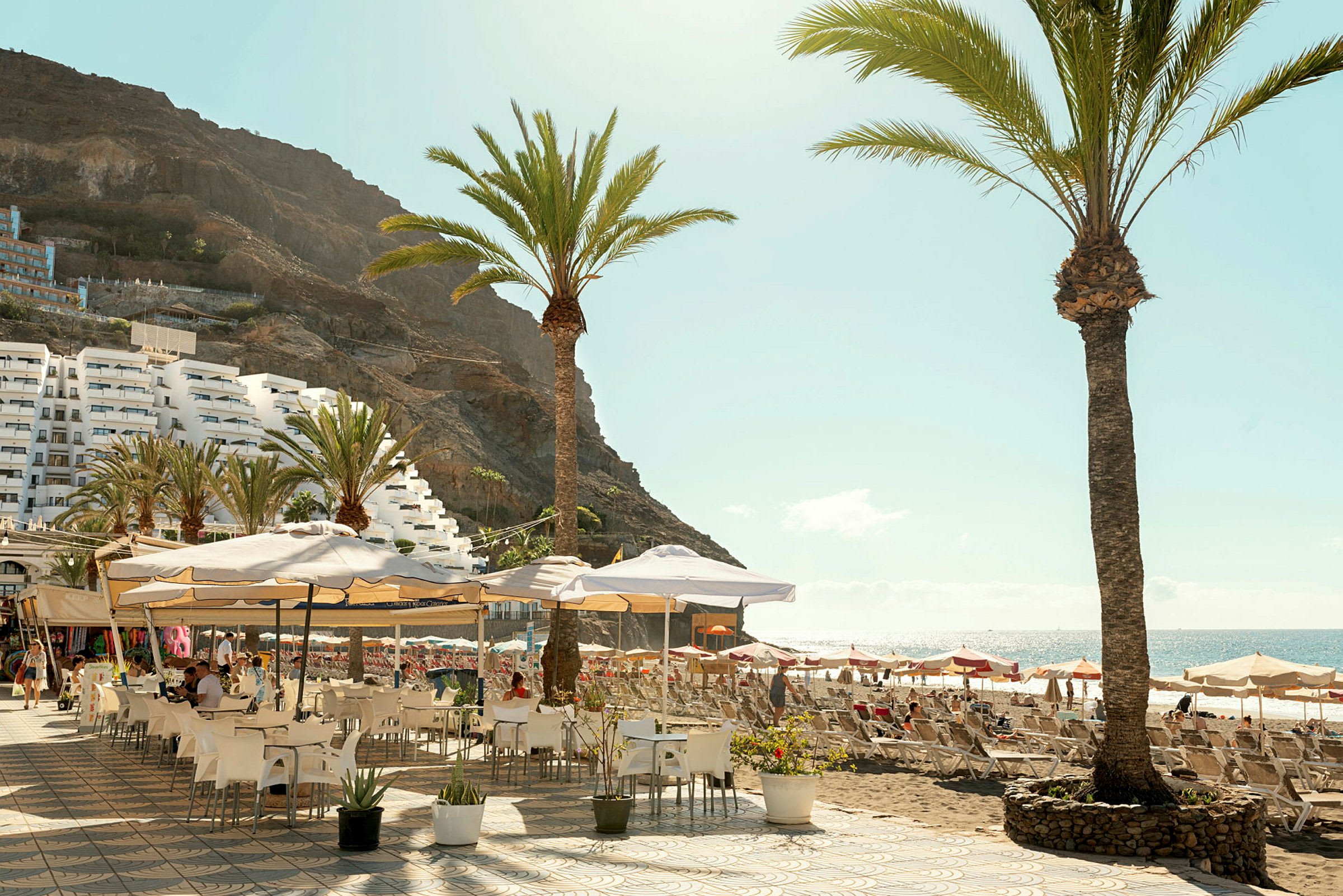 Utendørsrestaurant med parasoller og palmer nær en strand. (AI-generert tekst)