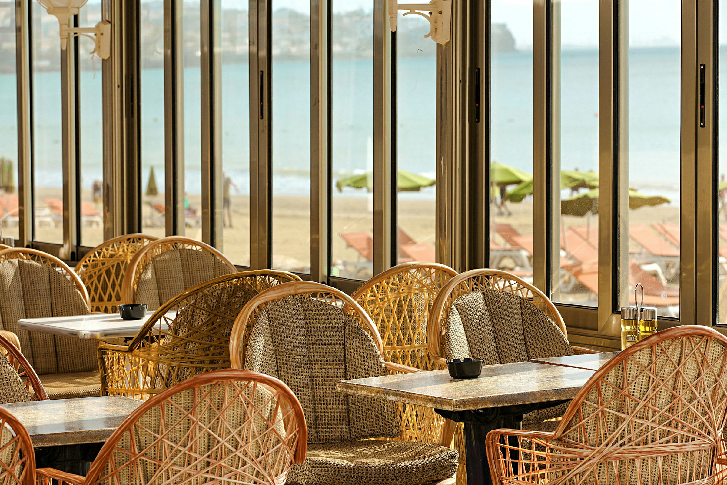 Utsikt fra en restaurant med kurvstoler og bord samt panoramavinduer med utsikt over stranden og sjøen. (AI-generert tekst)
