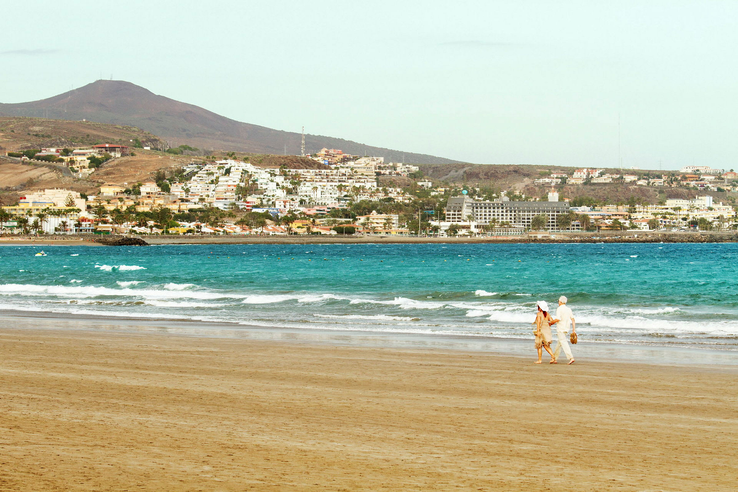 Et par går på en sandstrand med havet og en by i bakgrunnen. (AI-generert tekst)