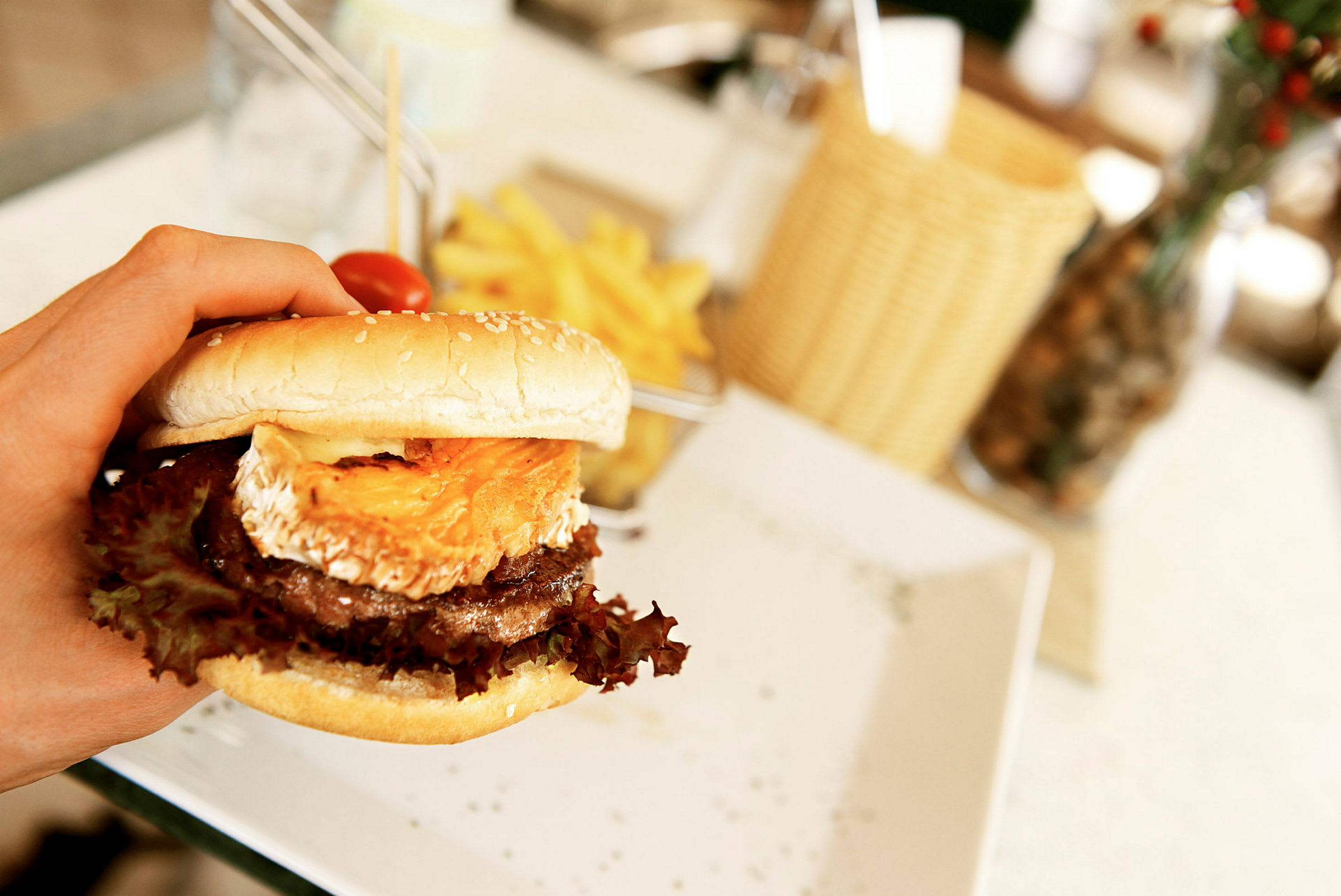 En hamburger med ost og salat holdes i en hånd, med pommes frites på en tallerken i bakgrunnen. (AI-generert tekst)