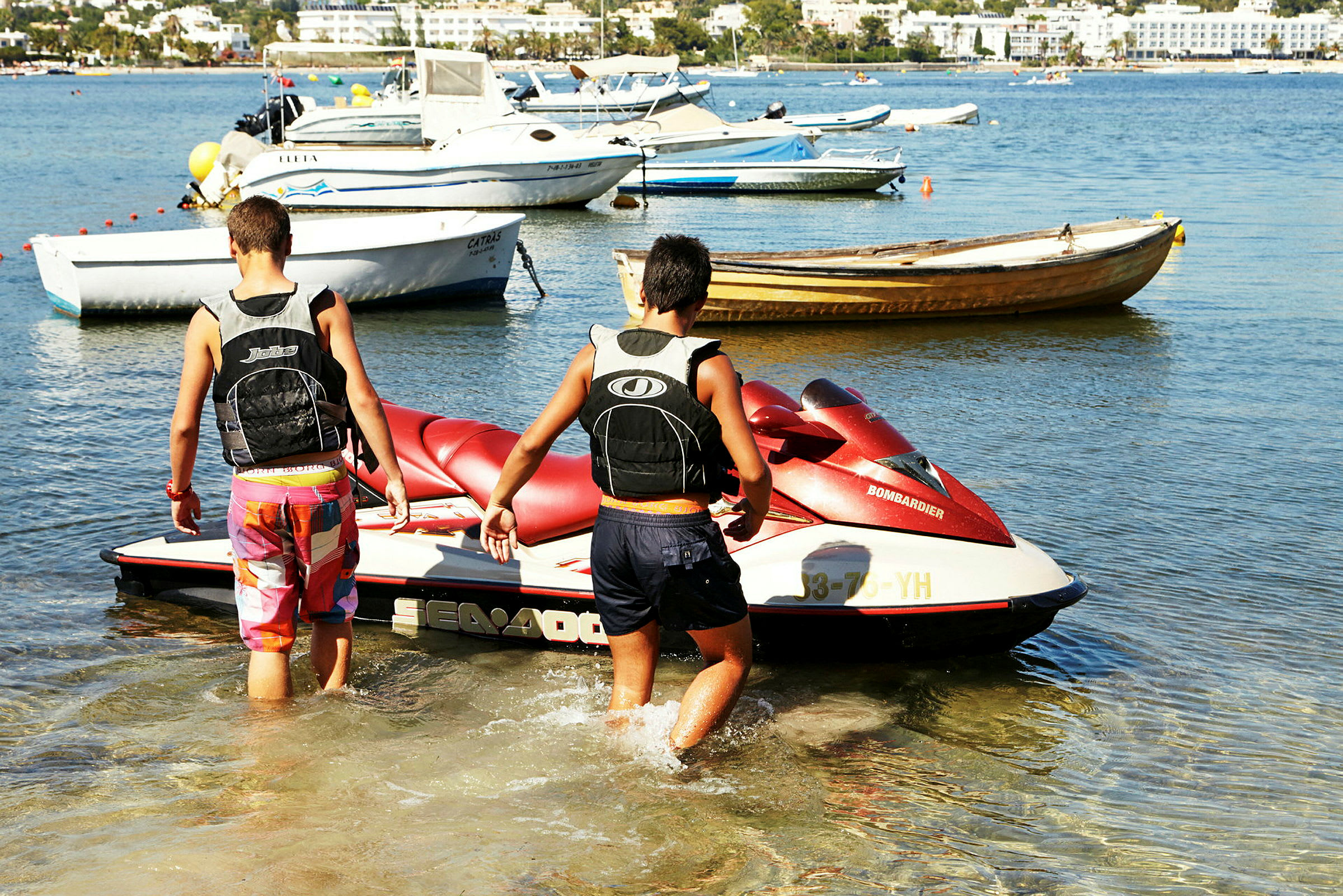 To personer trekker en vannscooter ut i vannet på en strand, med flere båter forankret i bakgrunnen. (AI-generert tekst)
