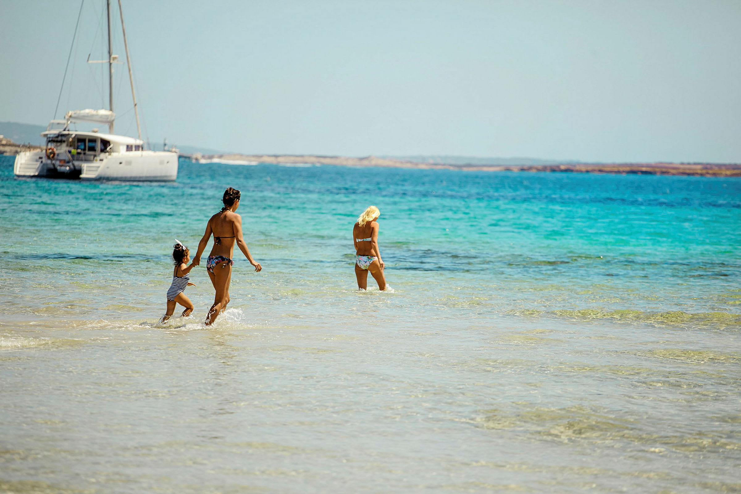 Tre personer går i grunt, klart vann på en strand, med en seilbåt i bakgrunnen. (AI-generert tekst)