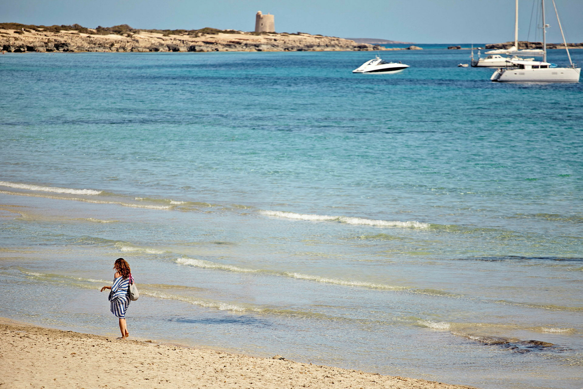 En person går langs en sandstrand ved havet med klare bølger. I bakgrunnen sees en liten båt og en seilbåt ute på vannet samt en klippe med en bygning. (AI-generert tekst)