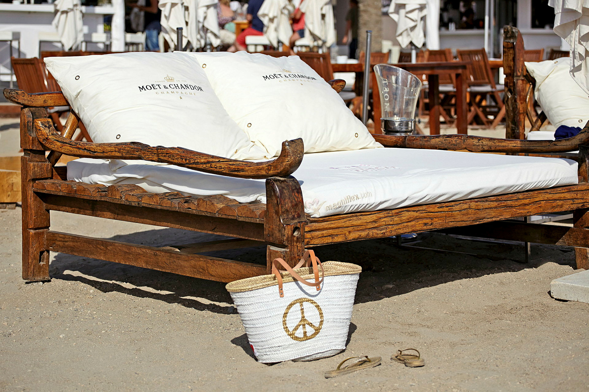 Bilde som viser en trebenk eller sofa med hvite puter merket 'Moët & Chandon' og en hvit strandveske med et gyldent fredstegn som står i sanden. I bakgrunnen sees utemøbler og parasoller. (AI-generert tekst)