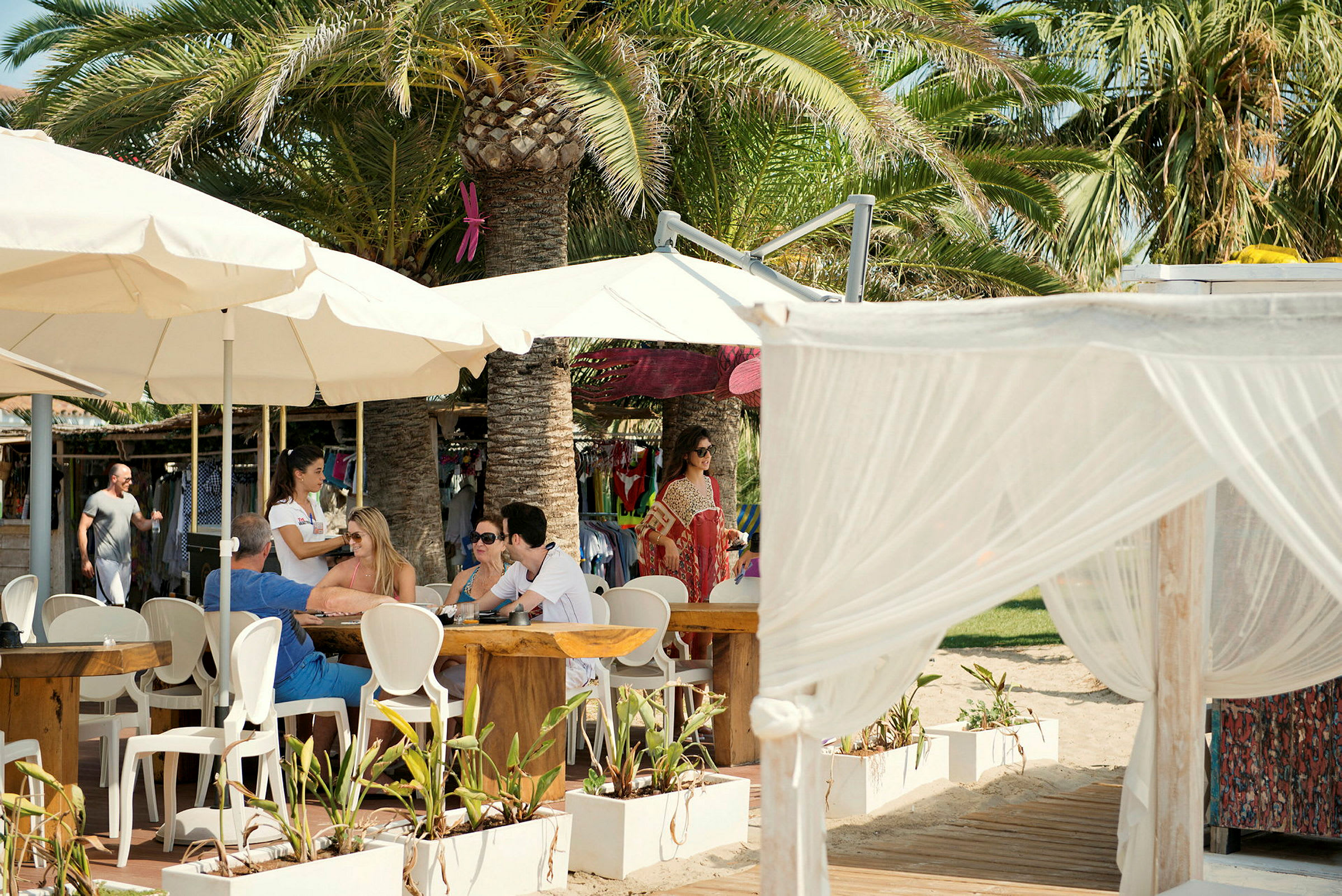 En uterestaurant med bord og stoler under parasoller ved en strandmiljø, omgitt av palmer og tekstilterrasser. (AI-generert tekst)