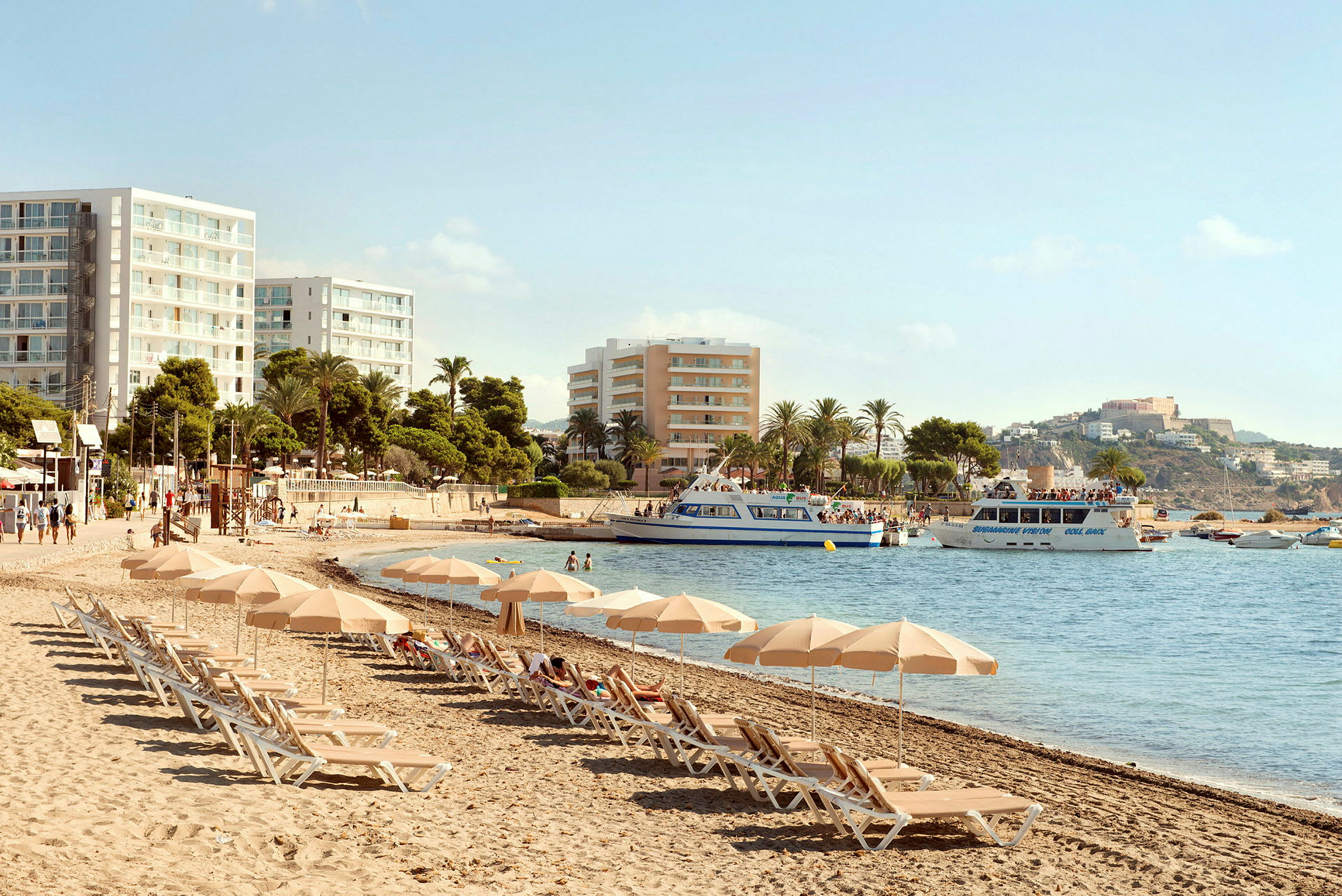 Strand med solsenger og parasoller, hotellbygninger i bakgrunnen og båter på vannet. (AI-generert tekst)