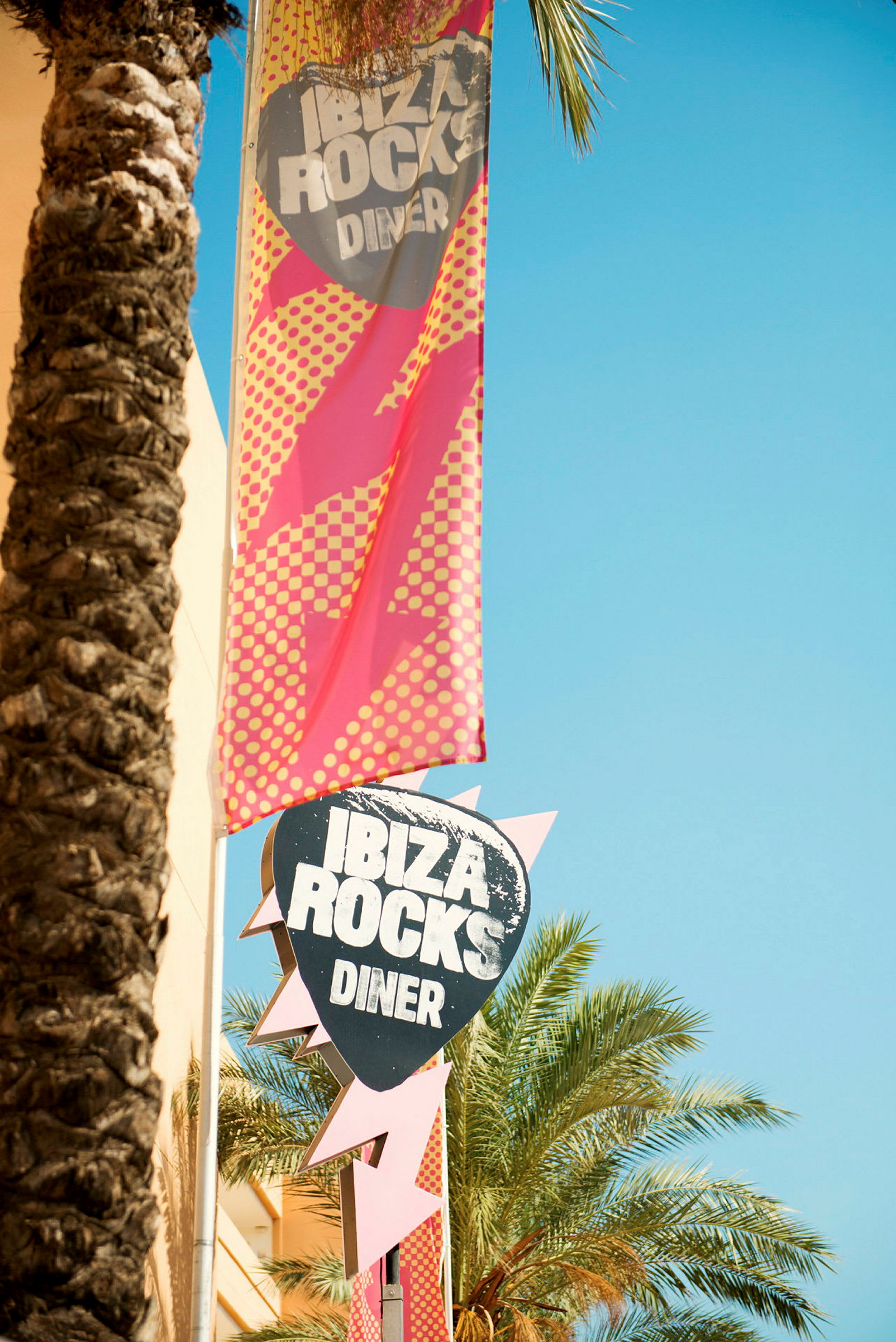 Et skilt og banner for Ibiza Rocks Diner, omgitt av palmer og klar himmel. (AI-generert tekst)