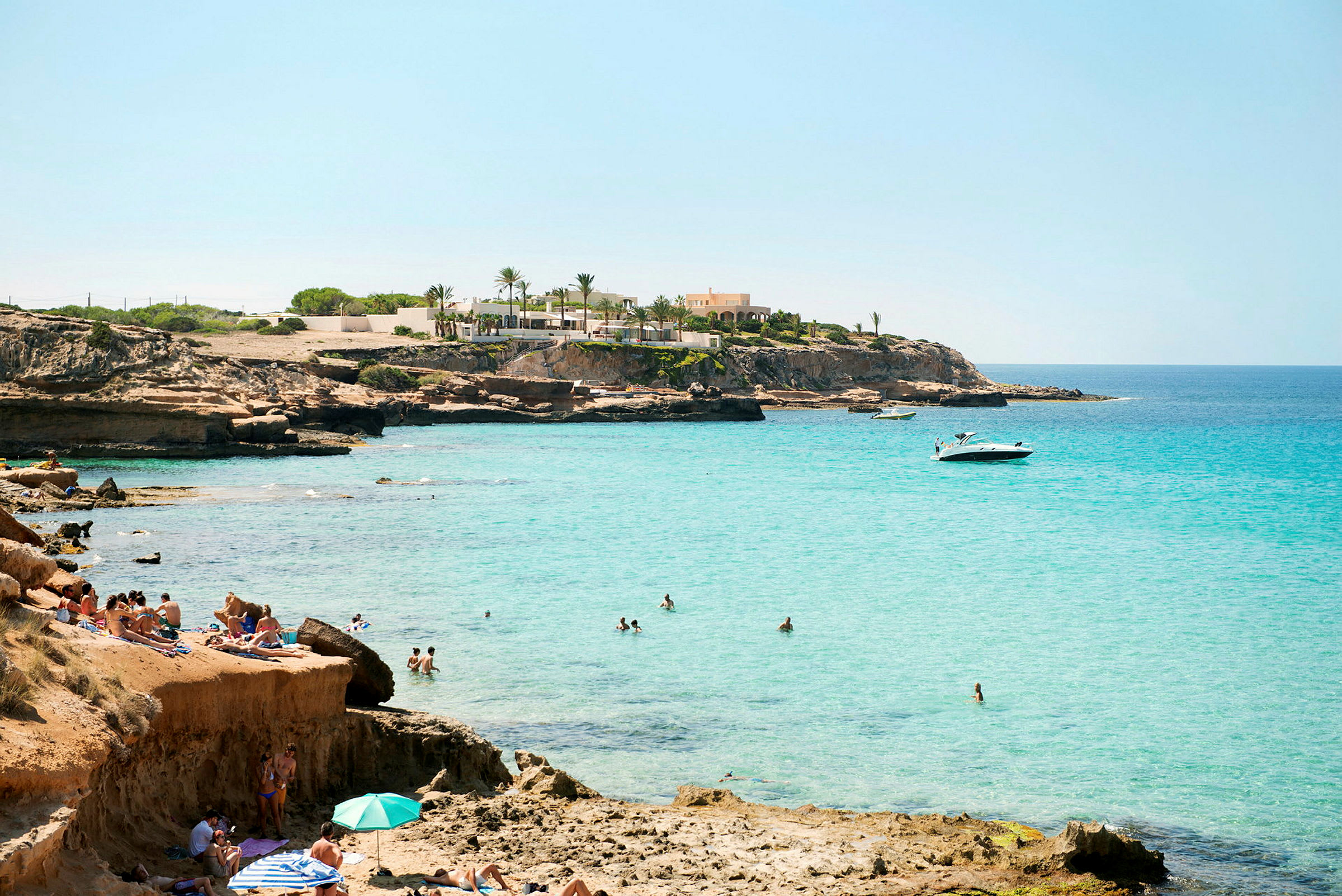 En klippefylt strand med turkisblått vann og folk som bader og soler seg. (AI-generert tekst)