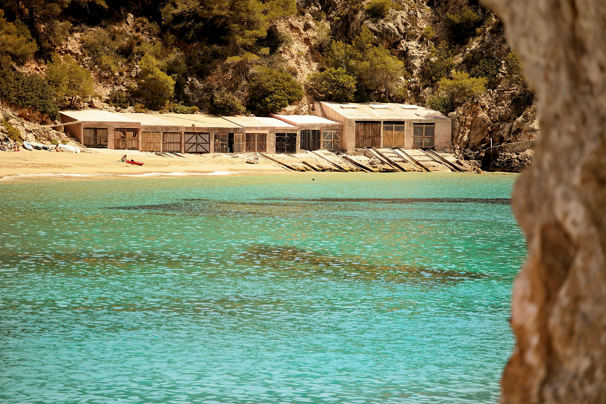 En liten strand med klart, turkist vann, trehytter og grønne trær ved en klippevegg. (AI-generert tekst)