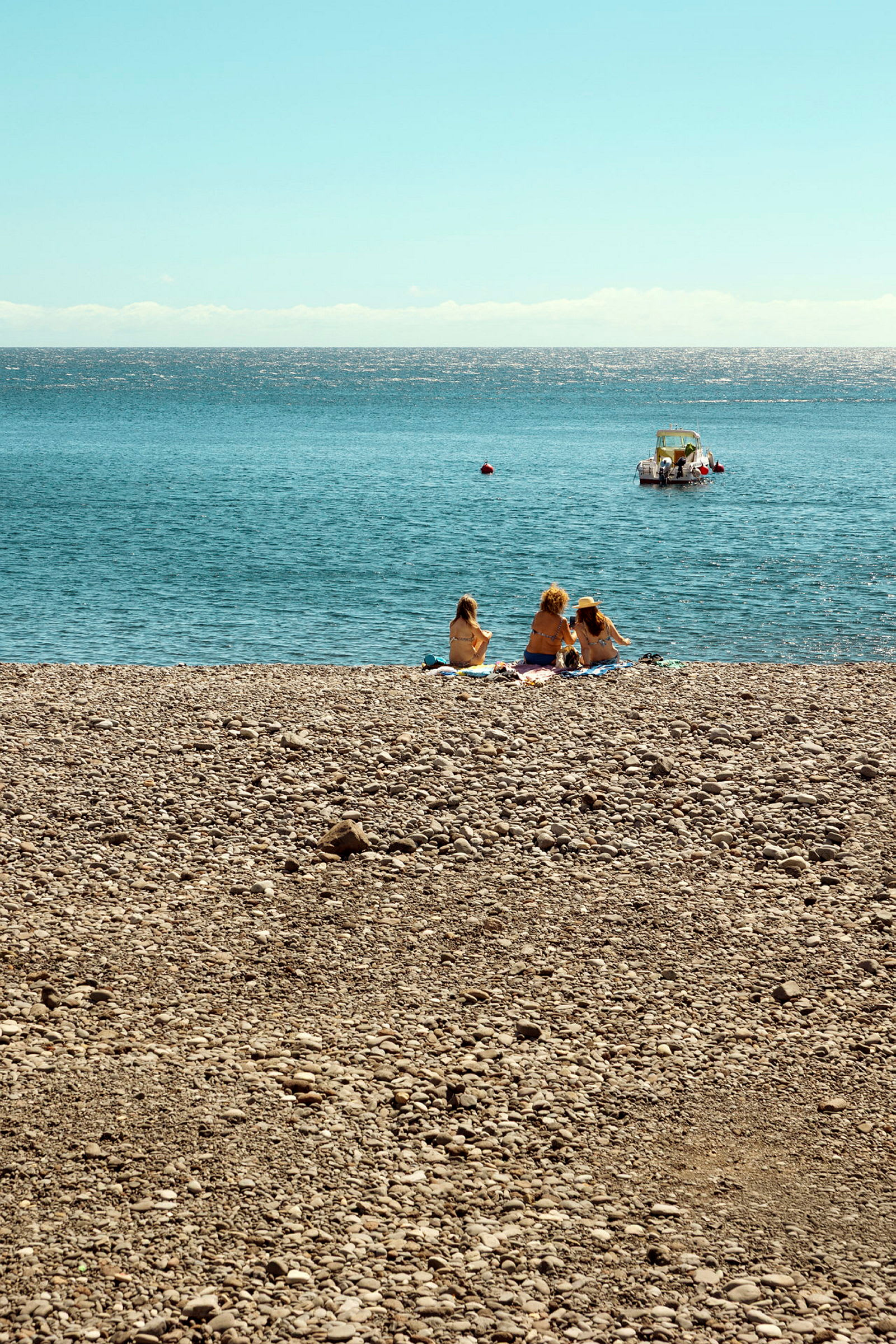Tre personer sitter på en steinstrand med havet i bakgrunnen og en båt lenger ute på vannet. (AI-generert tekst)