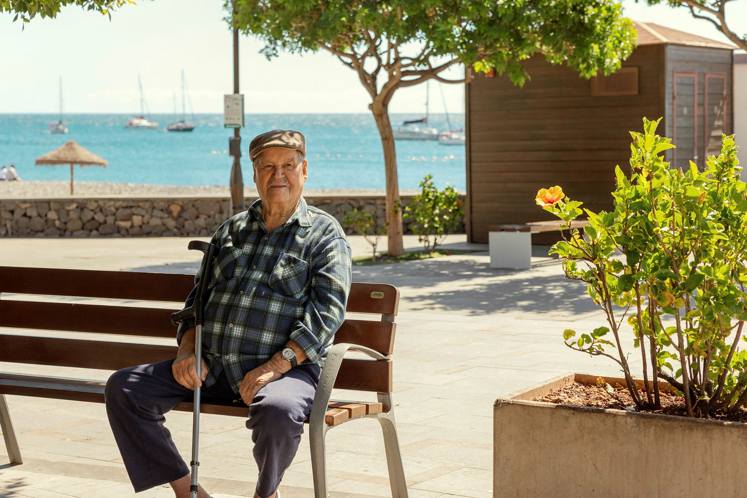 Person sitter på en trebänk ved en strandpromenade med havet og seilbåter i bakgrunnen. (AI-generert tekst)