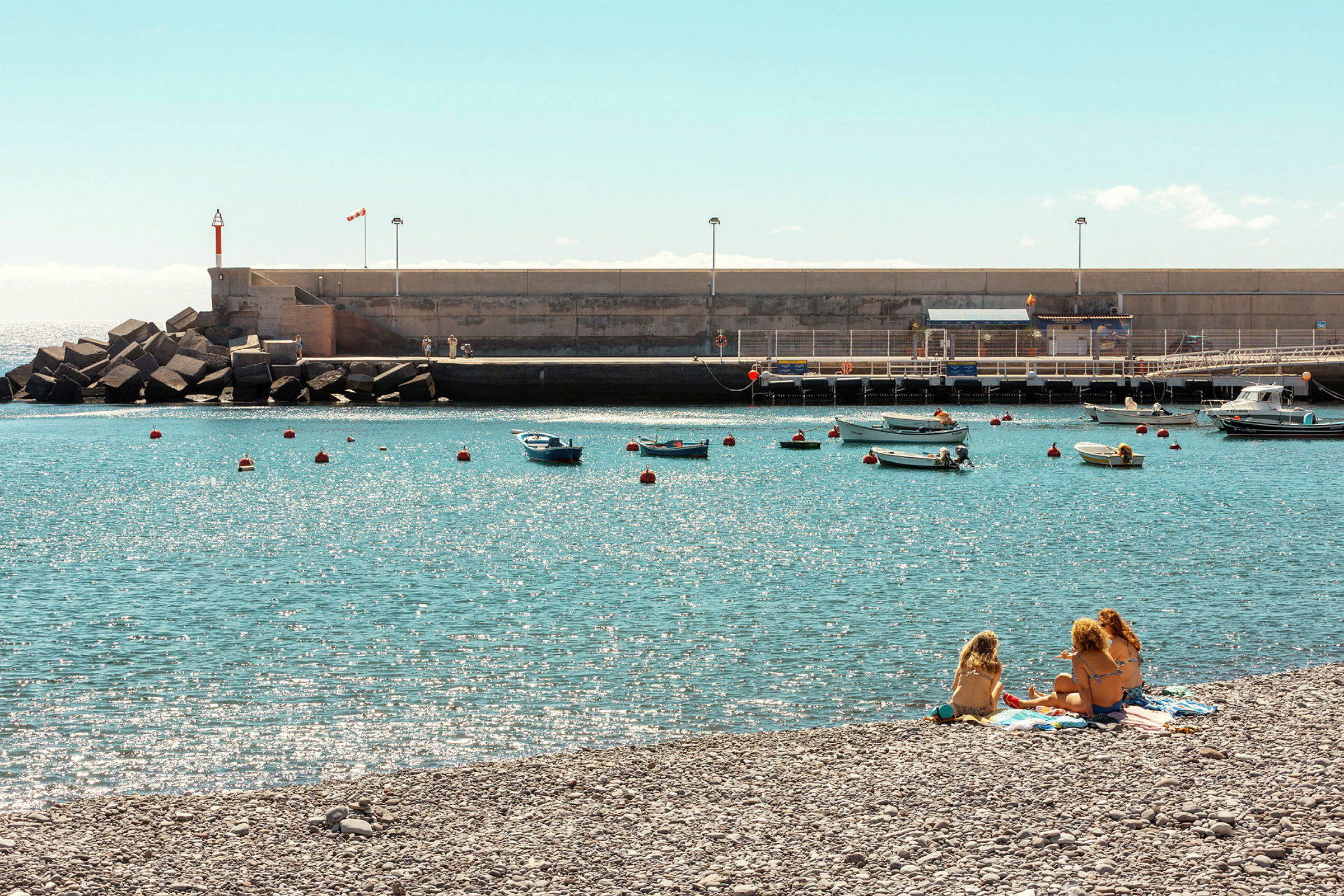 En strand med personer som sitter på håndklær ved vannet, med små båter fortøyd i havnen. (AI-generert tekst)