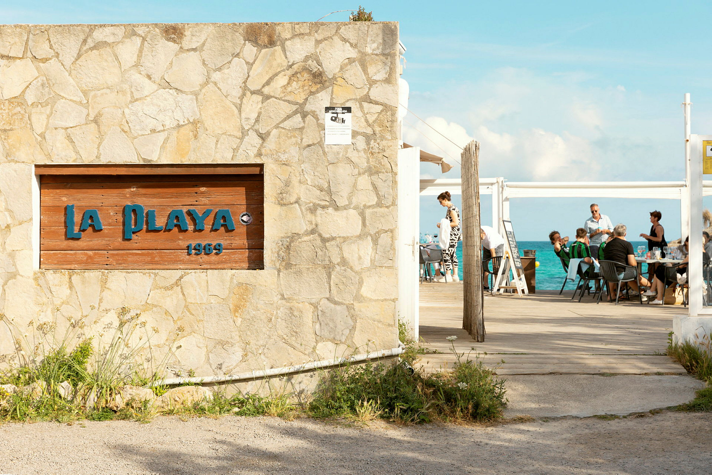 En steinmur med et skilt som har teksten 'La Playa 1969' foran en uteservering med havet i bakgrunnen. (AI-generert tekst)