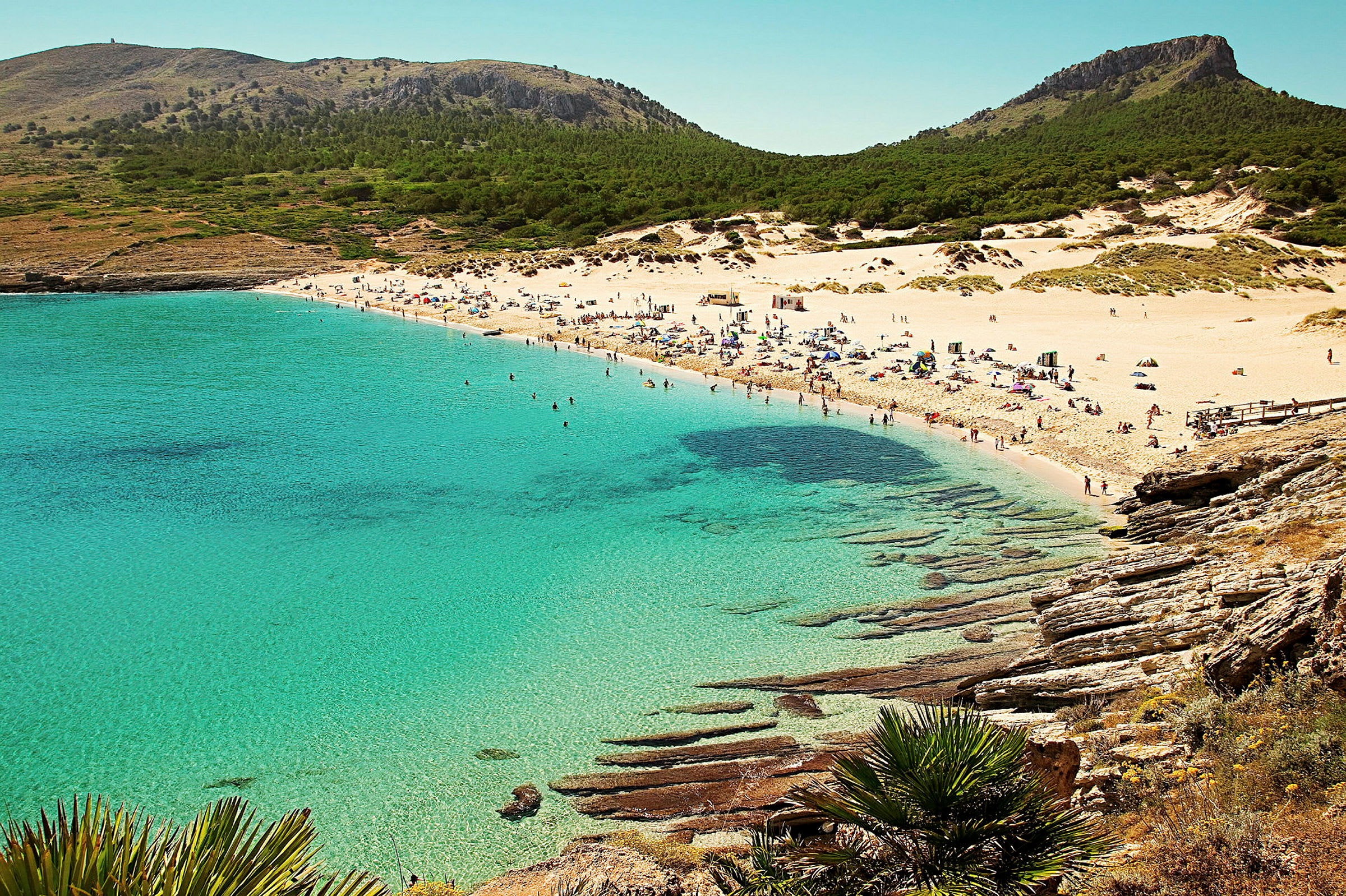 En sandstrand med krystallklart vann og folk som soler seg og bader. Skogdekte fjell sees i bakgrunnen. (AI-generert tekst)