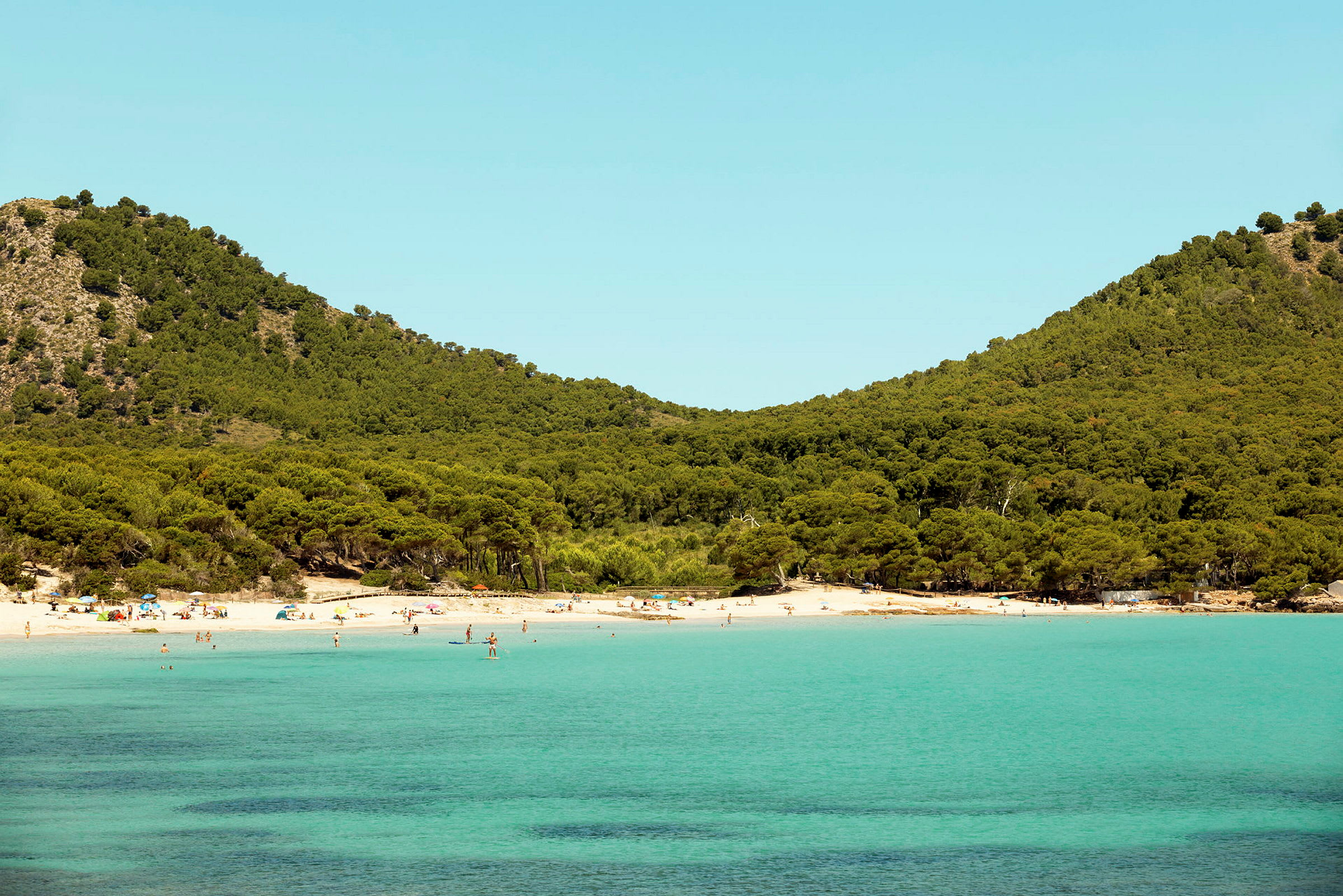 En strand med klart turkist vann og frodige grønne åser i bakgrunnen. (AI-generert tekst)