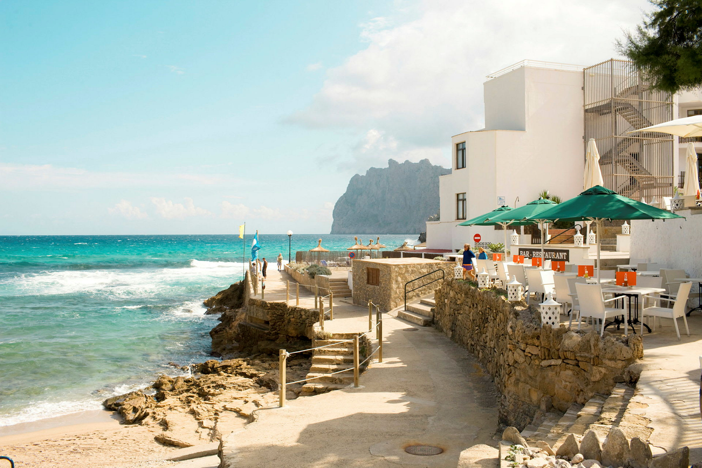 En steinete promenade ved havet nær en restaurant med uteservering. (AI-generert tekst)