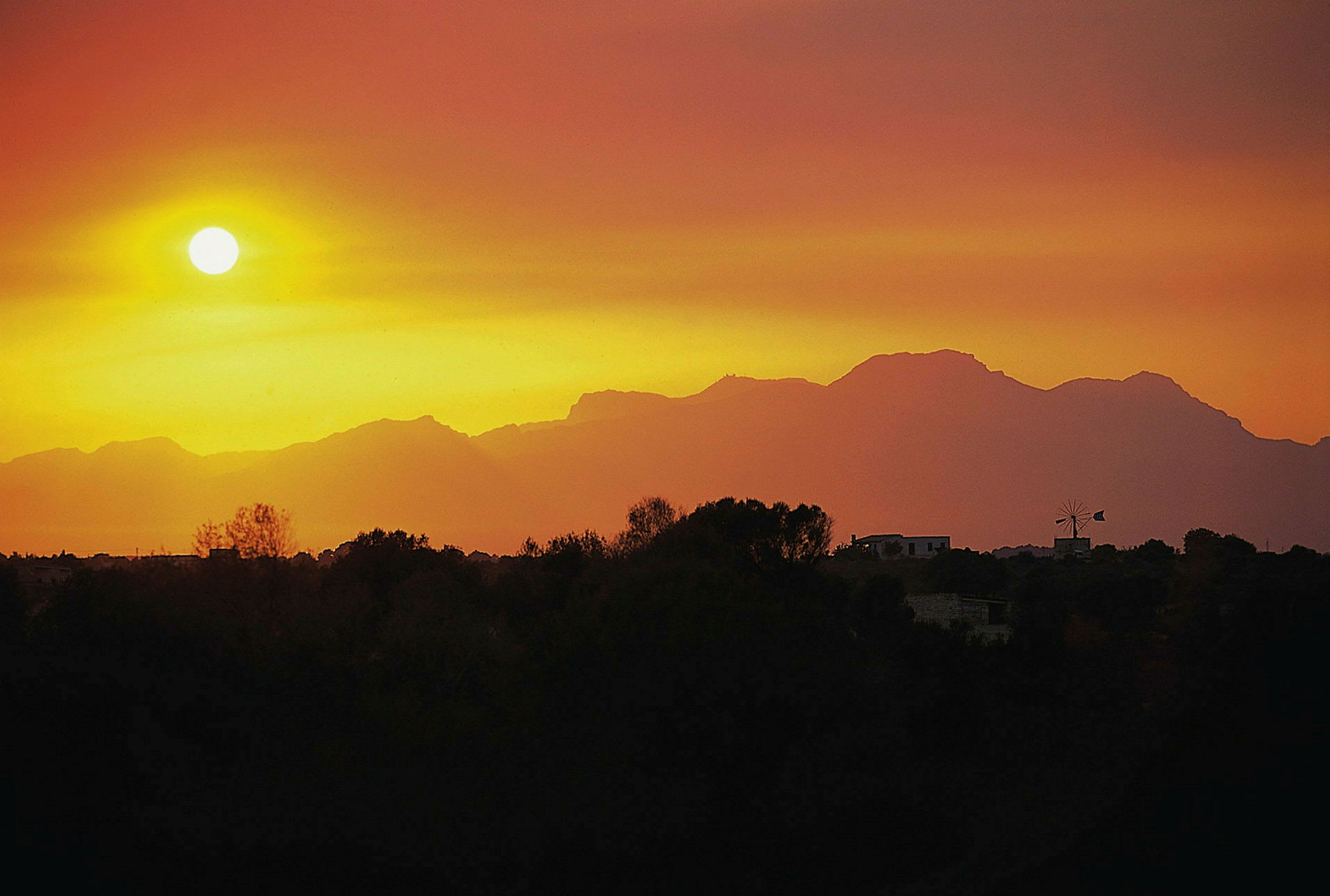 Solnedgang over silhuetten av et fjell med trær og bygninger i forgrunnen. (AI-generert tekst)