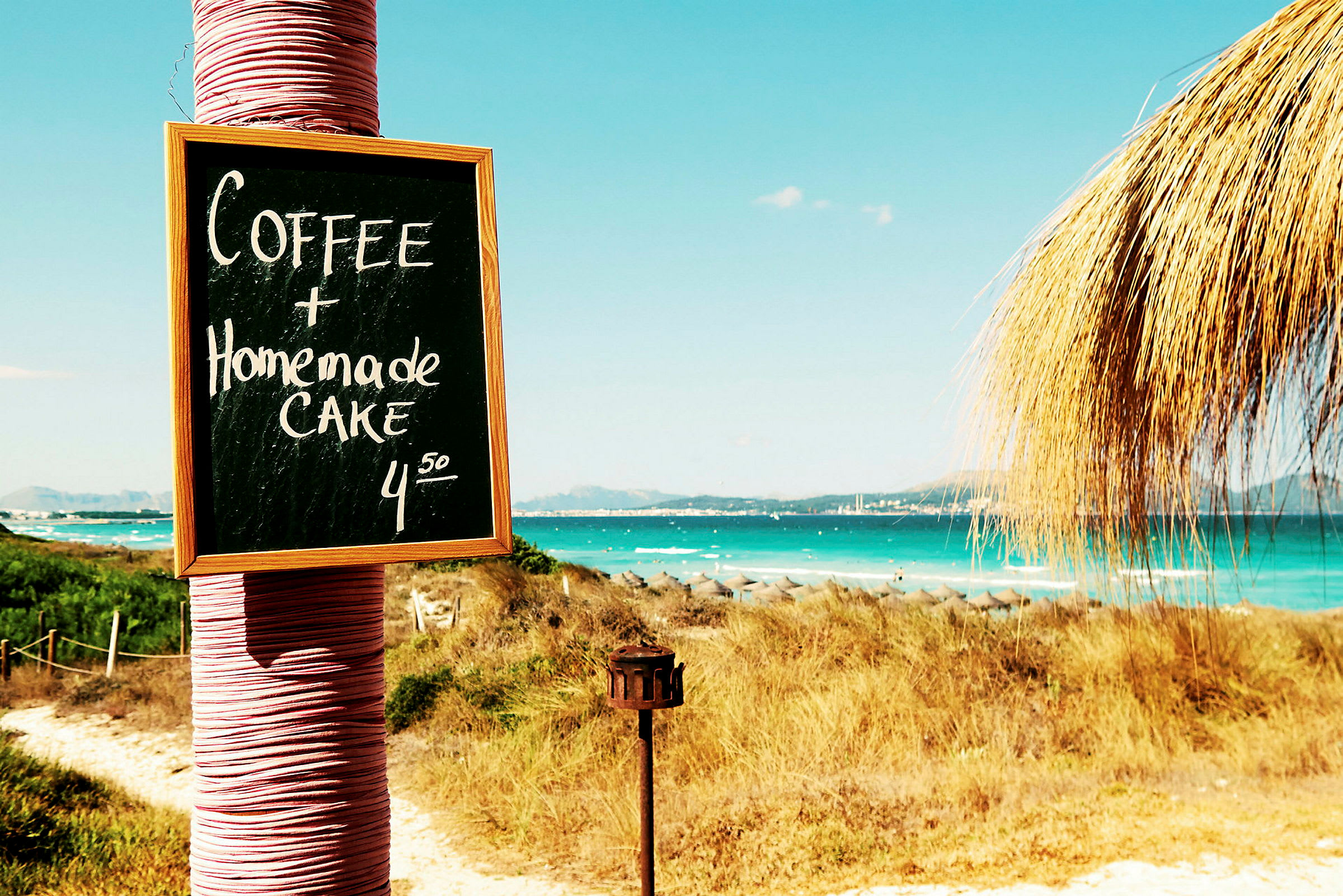 Skilt med teksten 'Coffee + Homemade Cake 4.50' ved stranden. (AI-generert tekst)
