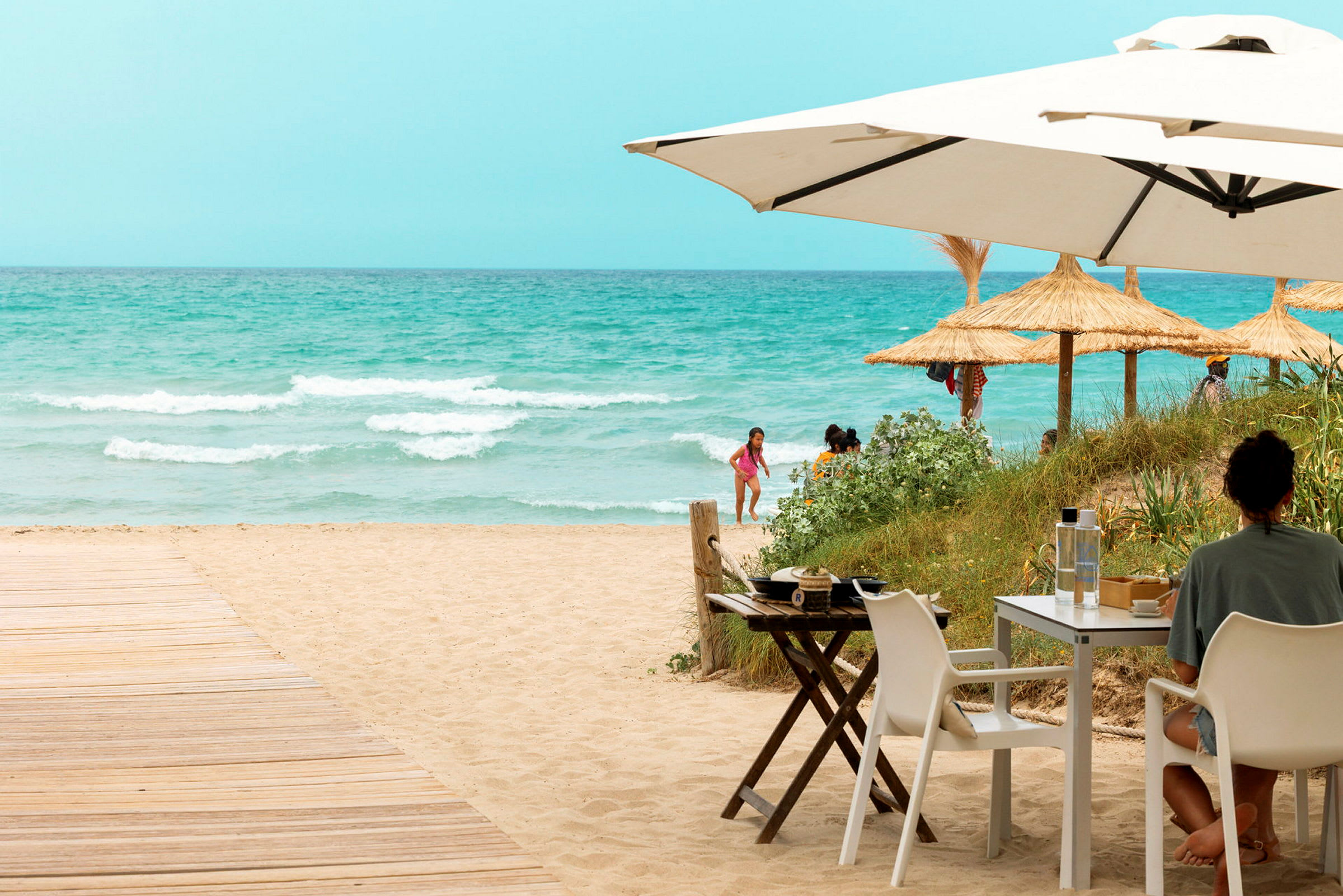 En person sitter ved et bord under en parasoll ved en sandstrand med hav og strandparasoller i bakgrunnen. (AI-generert tekst)