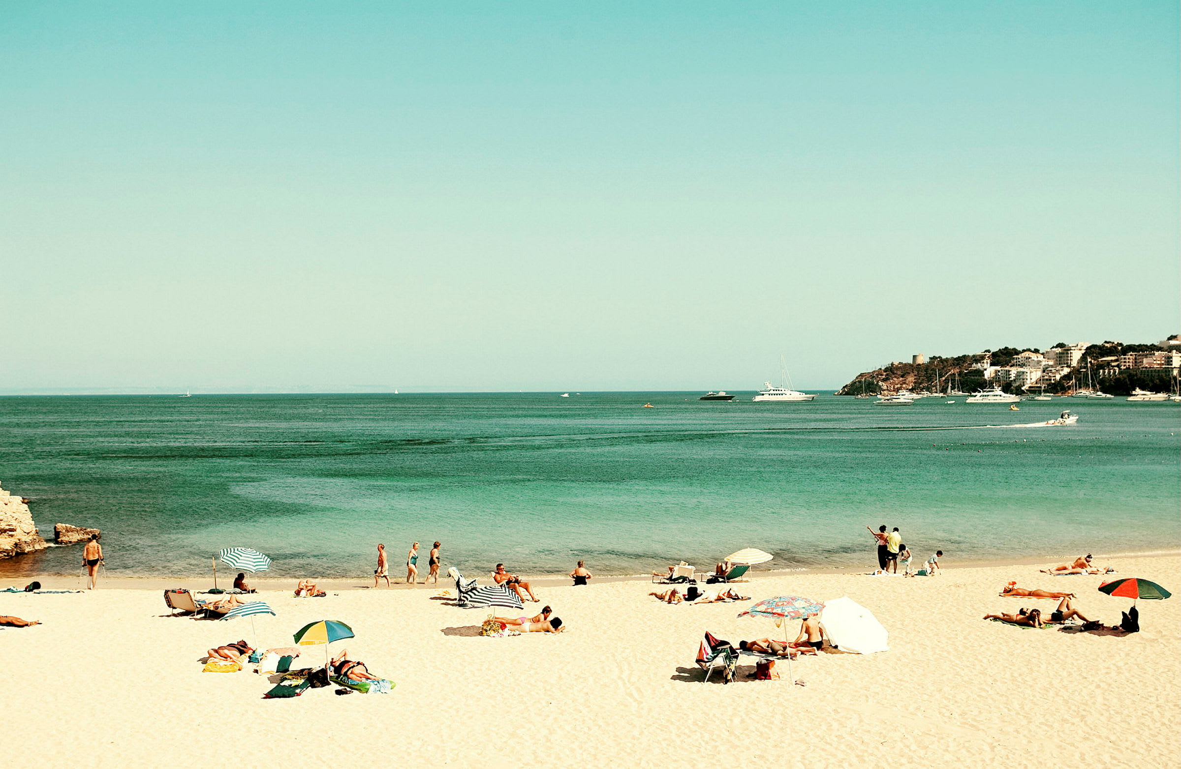 En strand med mennesker som soler seg og bader, parasoller og sjøen med båter i bakgrunnen. (AI-generert tekst)