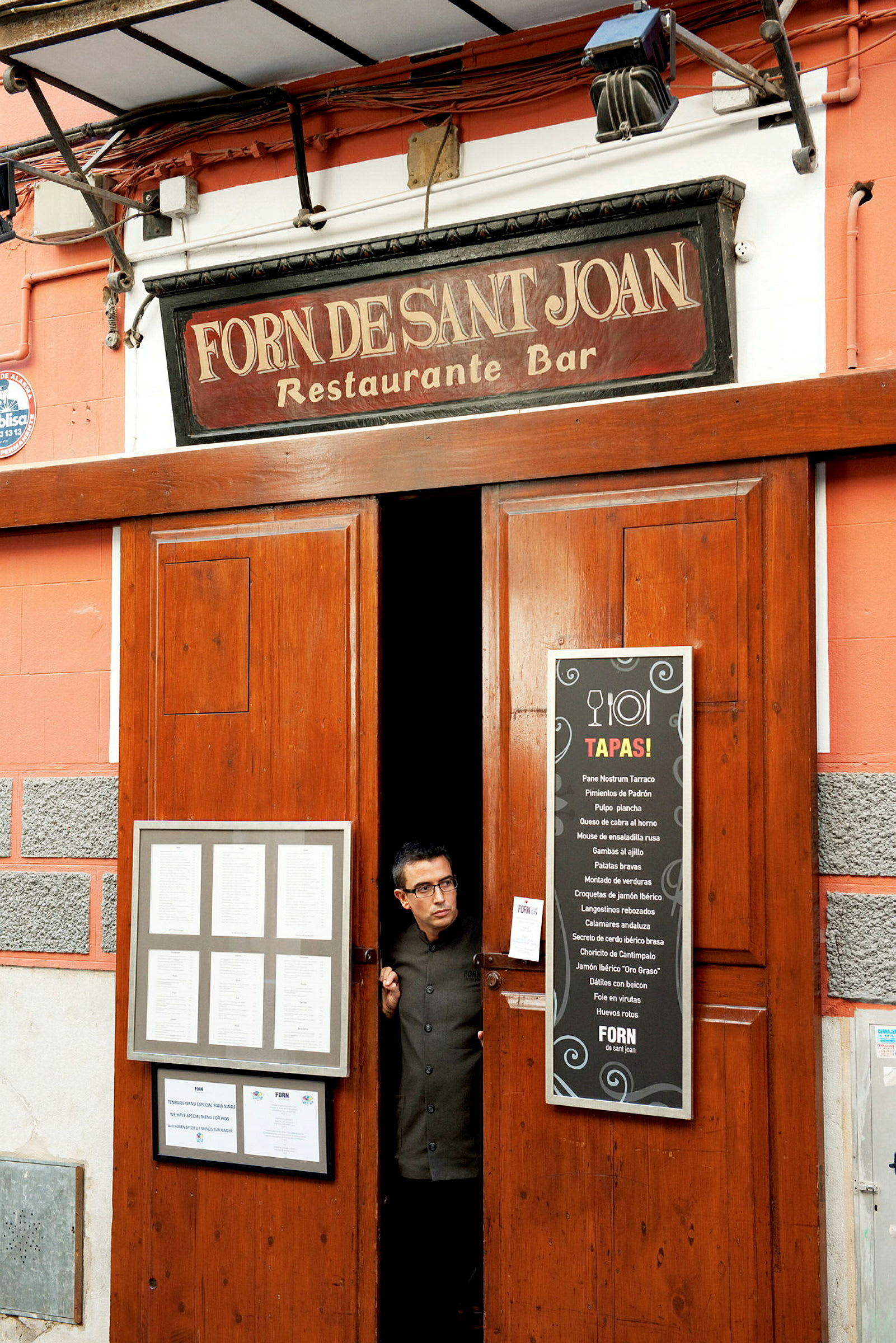 Inngangen til en restaurant og bar med et skilt som viser navnet 'Forn Desant Joan' og menyer utenfor. (AI-generert tekst)