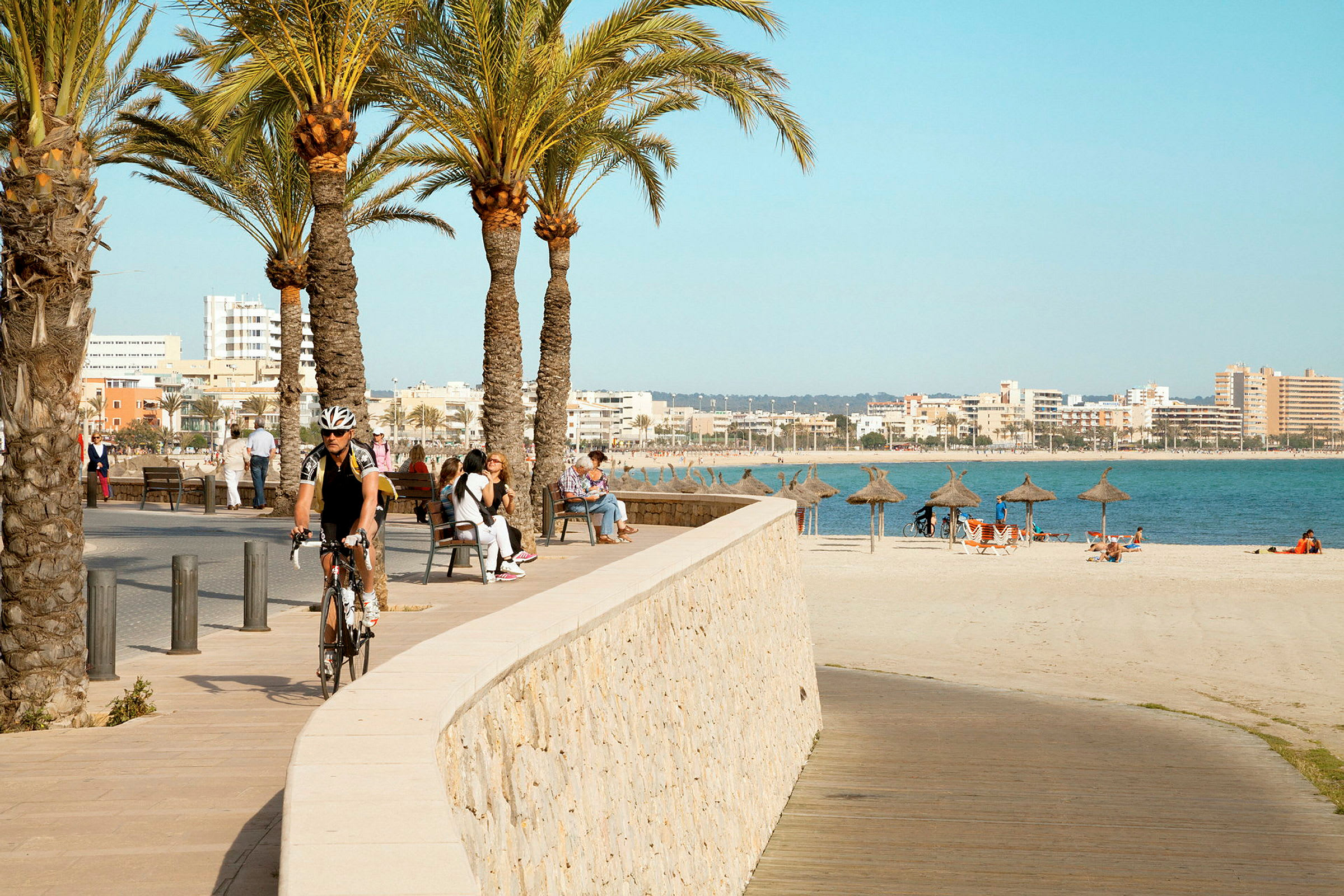 Strandpromenade med palmer ved havet, folk sitter på benker og solparasoller er plassert på stranden. (AI-generert tekst)