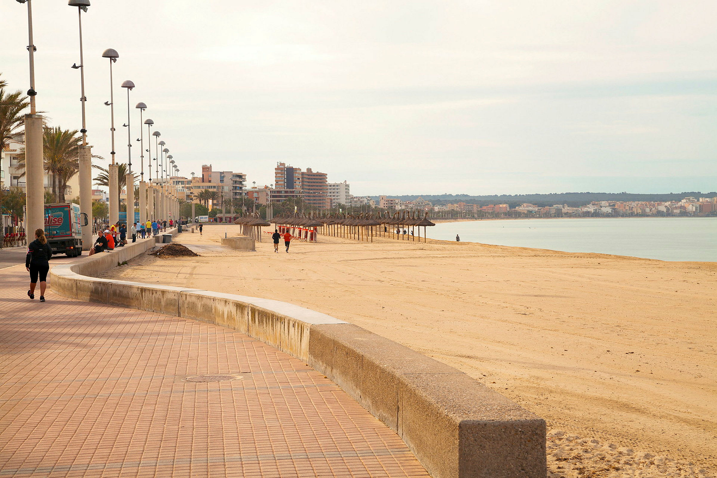 En bred strandpromenade med lyktestolper og mennesker som går langs sandstranden. Byen og havet er synlig i bakgrunnen. (AI-generert tekst)