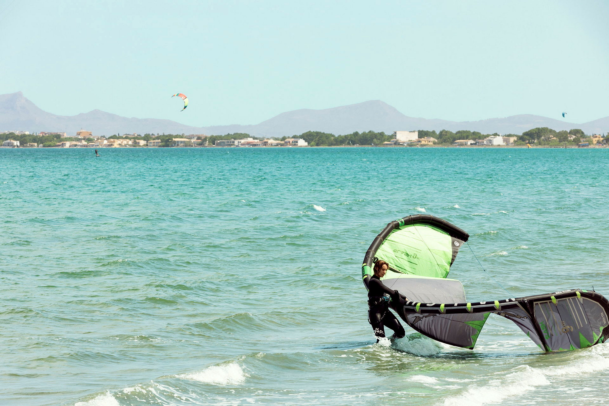 En person i vannet med en grønn og svart kitesurfing drage, med en by og fjell i bakgrunnen. (AI-generert tekst)