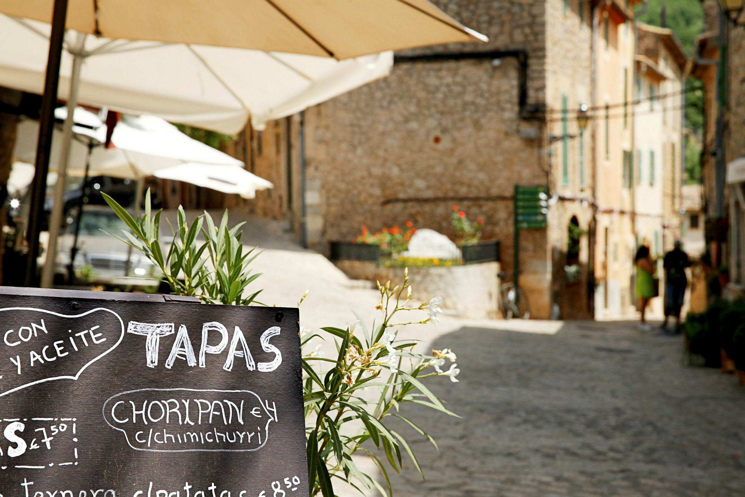 Gatebilde med en svart menytavle med teksten «tapas» i forgrunnen og parasoller samt steinhus i bakgrunnen. (AI-generert tekst)