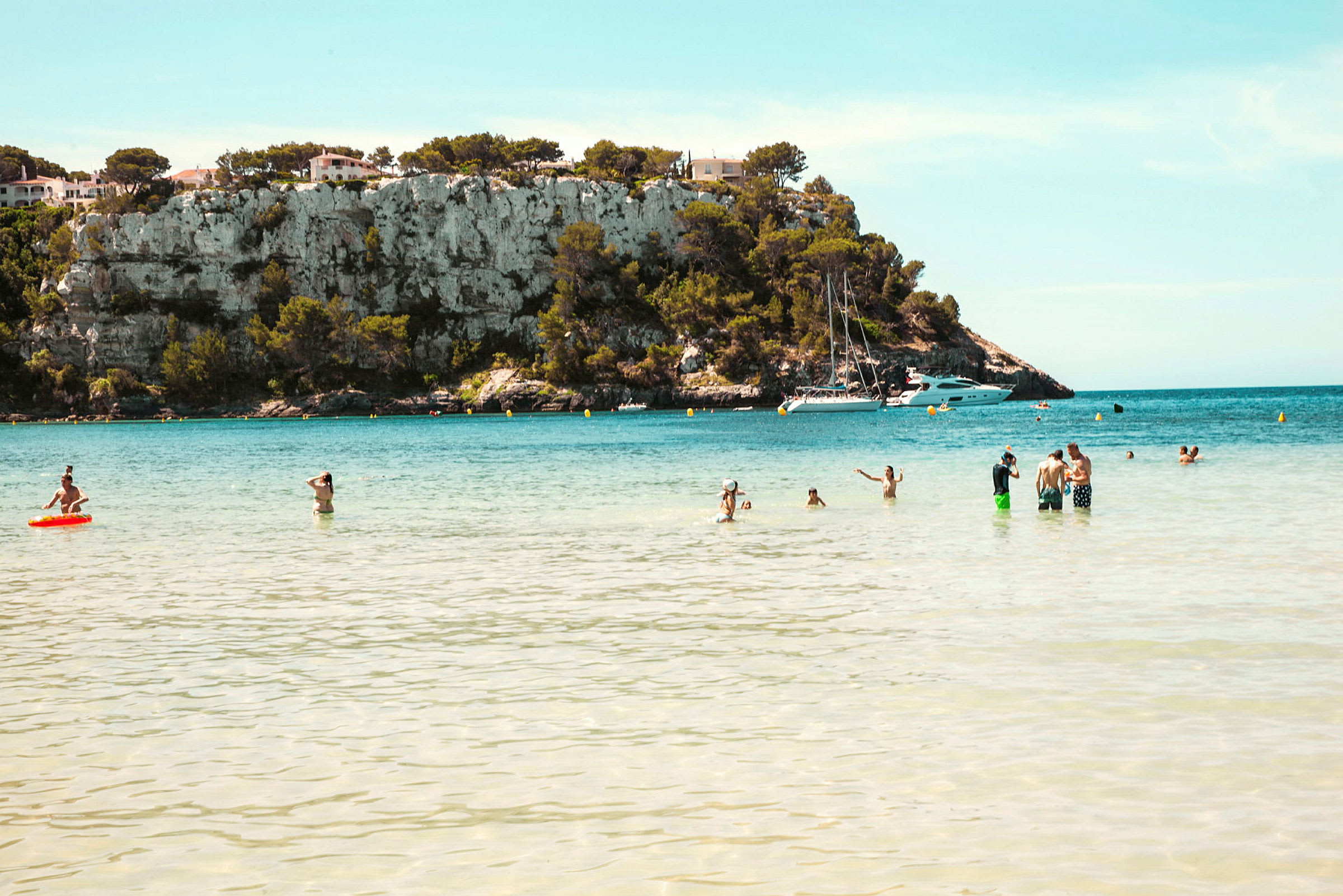 En strand med mennesker i vannet foran en ås med klipper og noen hus samt båter i bakgrunnen. (AI-generert tekst)