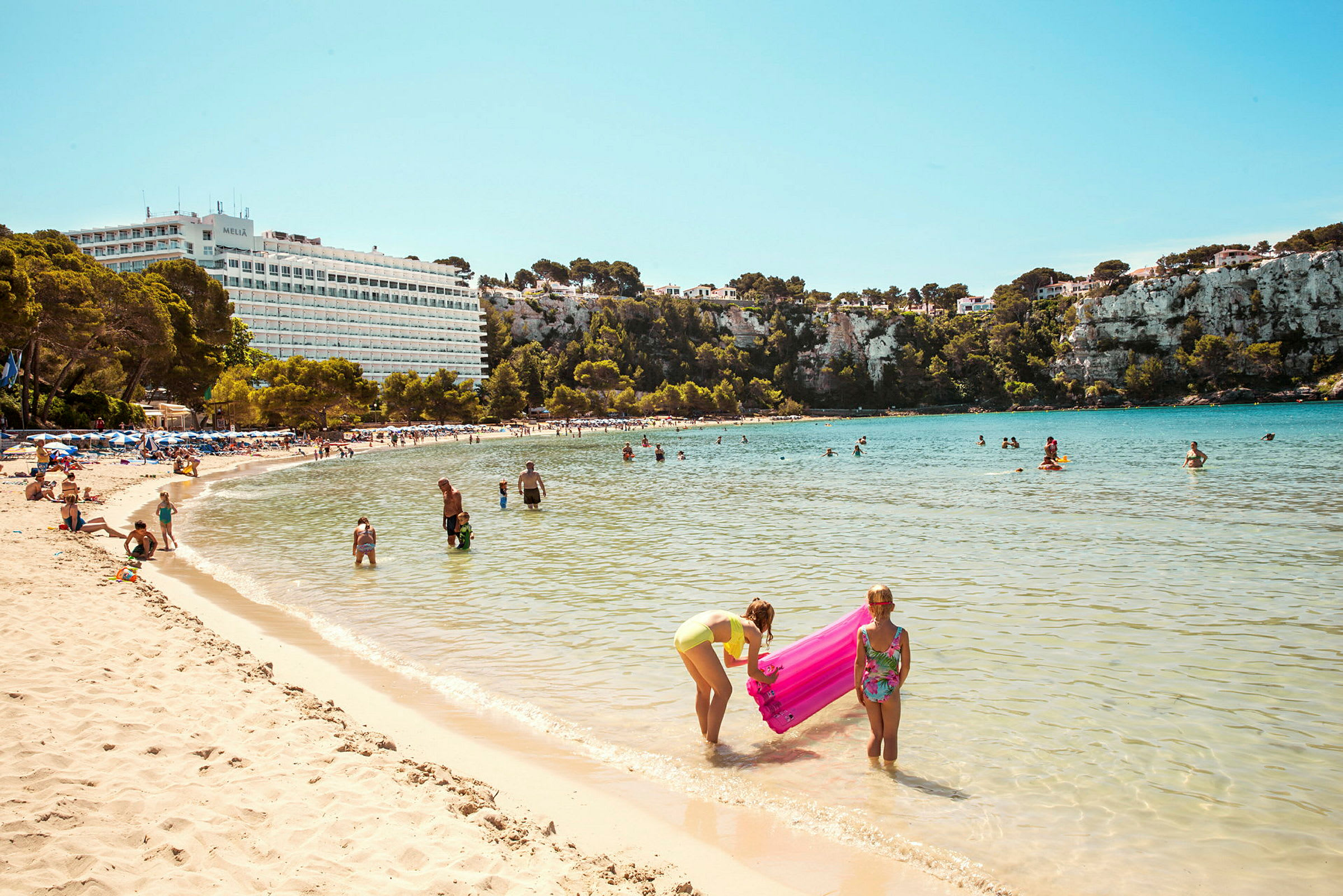Strand med folk som bader og leker i vannet foran en naturskjønn klippe og et hotell. (AI-generert tekst)