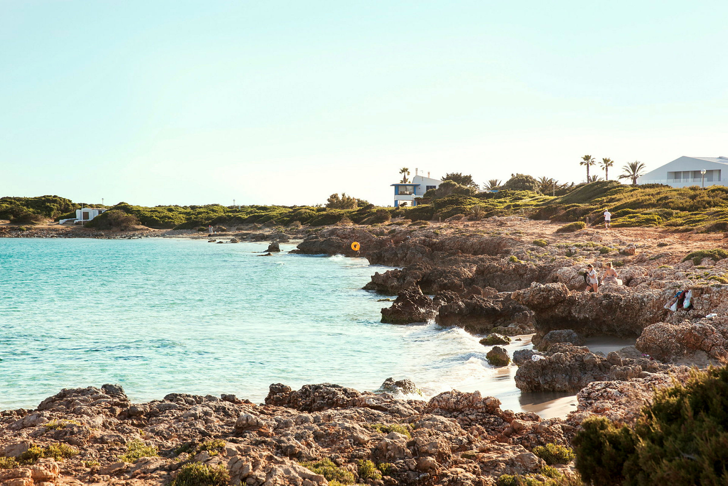 Strand med klart vann og steinete landskap, omkringliggende grøntområder og bygninger i bakgrunnen. (AI-generert tekst)