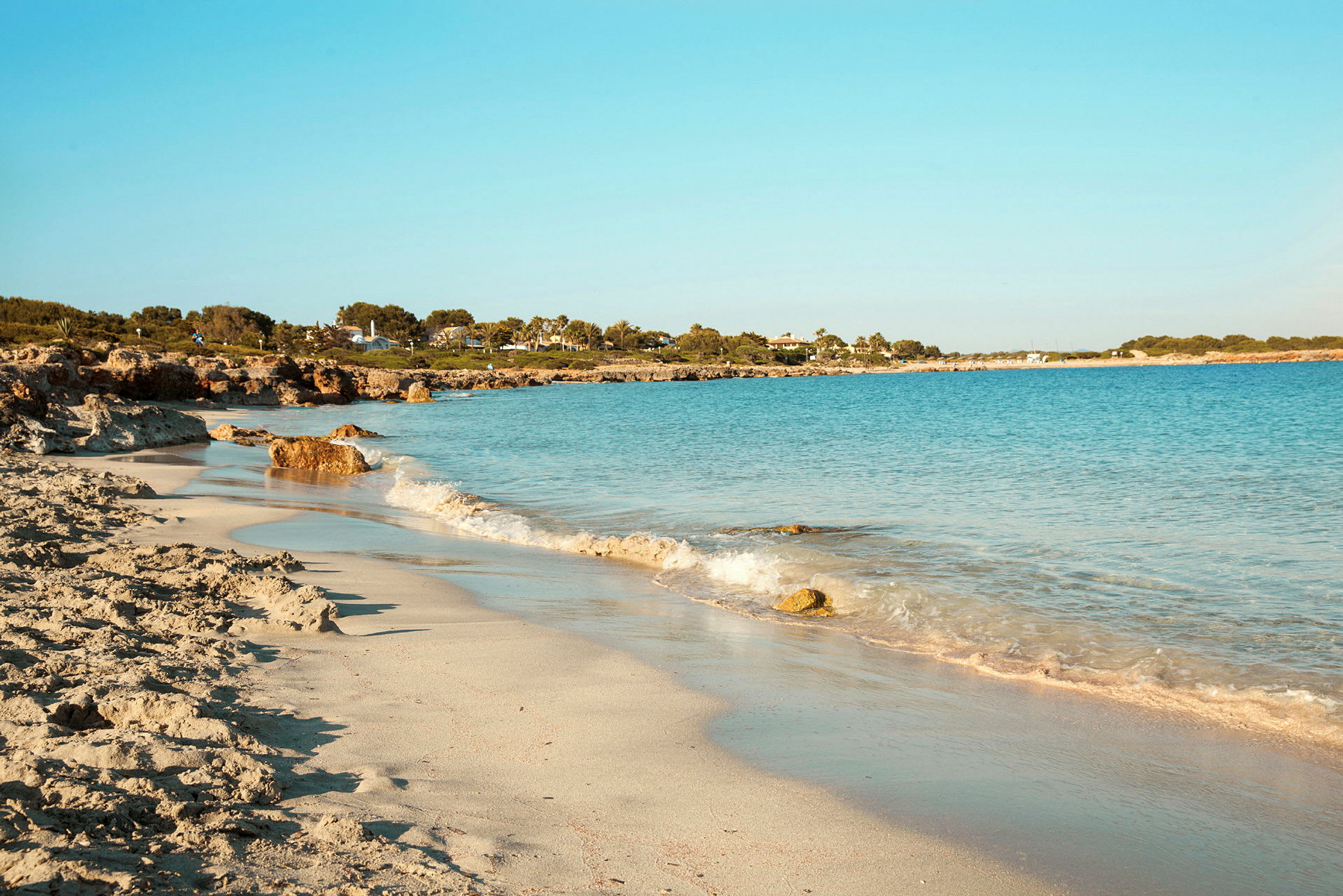 En strand med lys sand og blått hav, omgitt av grøntområder og bygninger i bakgrunnen. (AI-generert tekst)