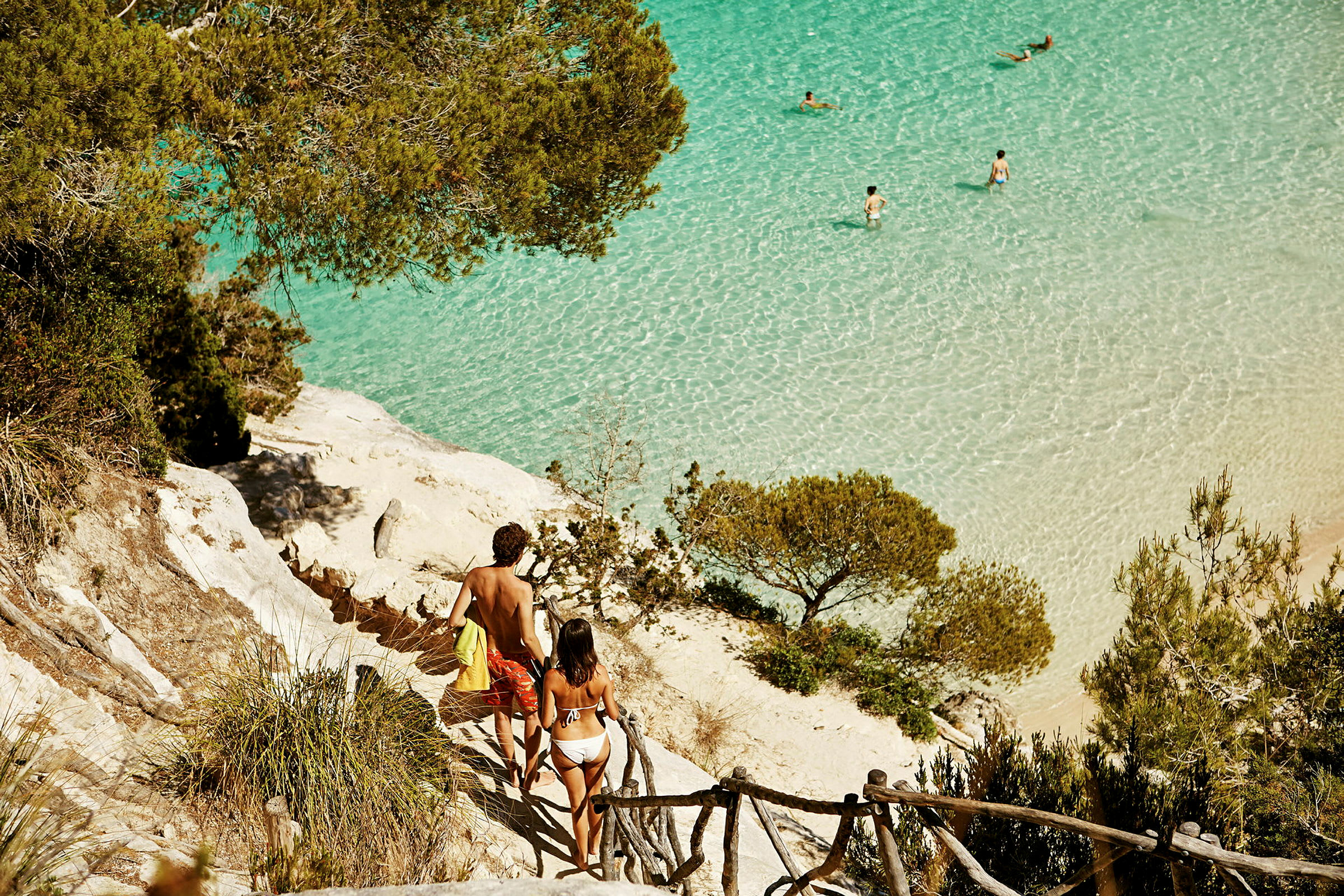 To personer går ned en steinete sti mot en strand med klart turkis vann, omgitt av vegetasjon. (AI-generert tekst)