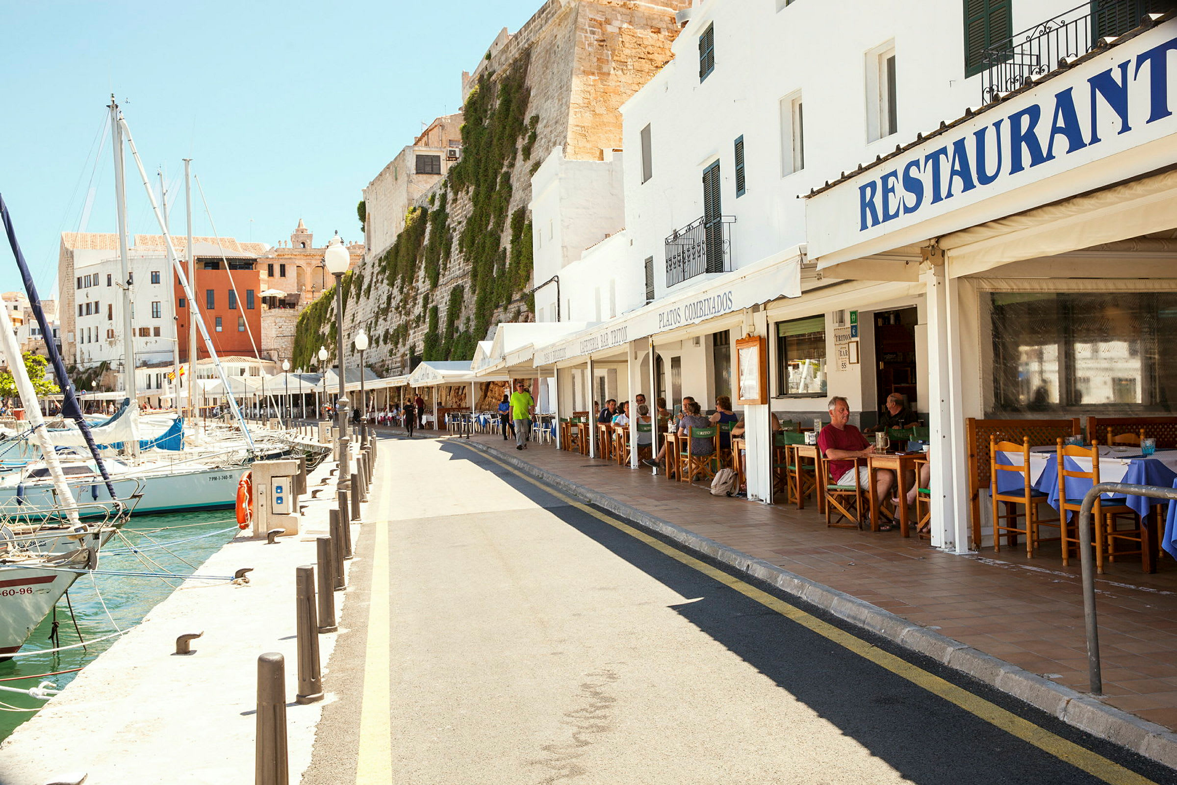 En havnepromenade med restauranter og båter ved bryggen. Det er en solfylt dag med folk som sitter ute på restaurantenes terrasser. (AI-generert tekst)