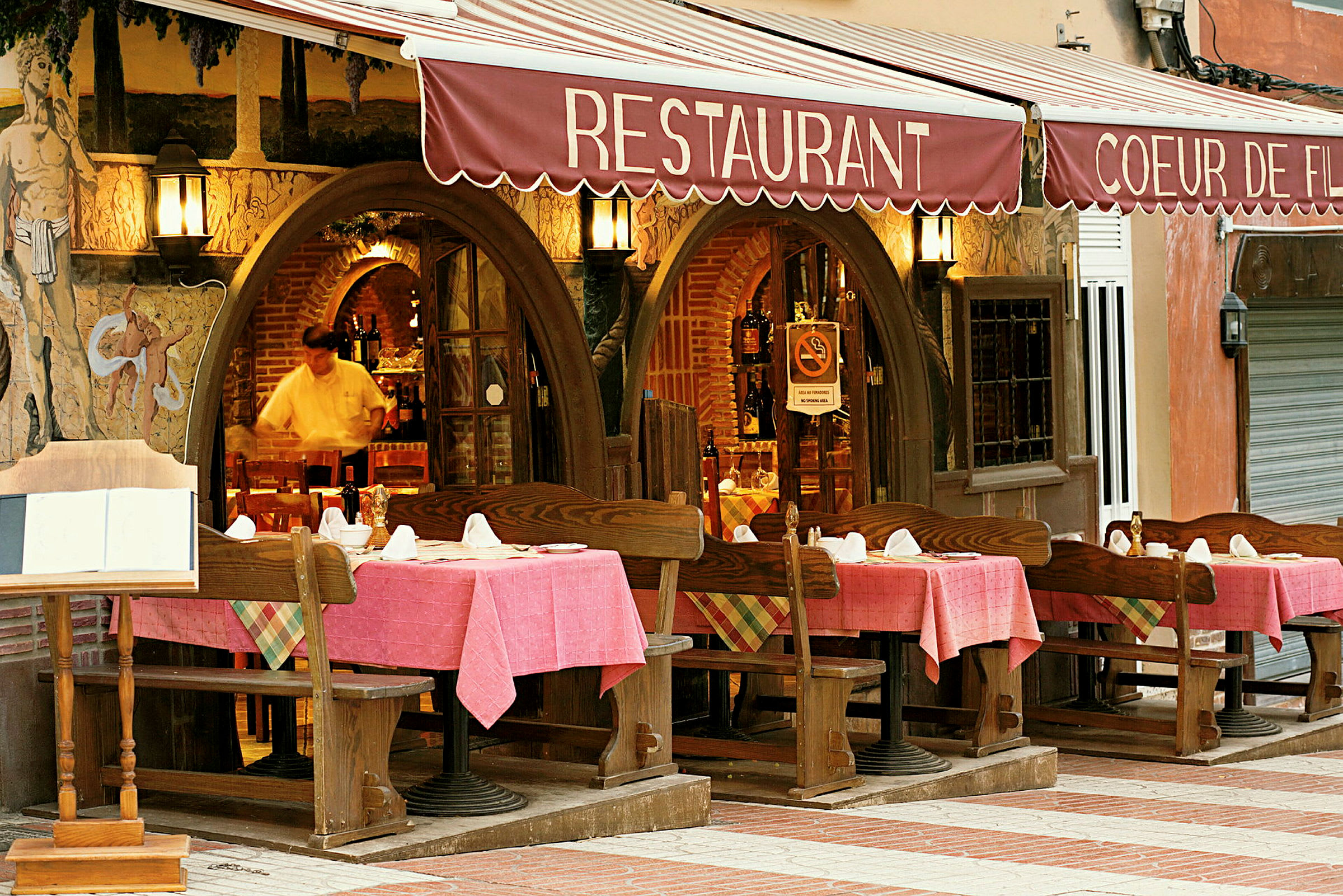 Inngangen til en restaurant med bord og stoler utendørs, dekket med rosa duker. (AI-generert tekst)