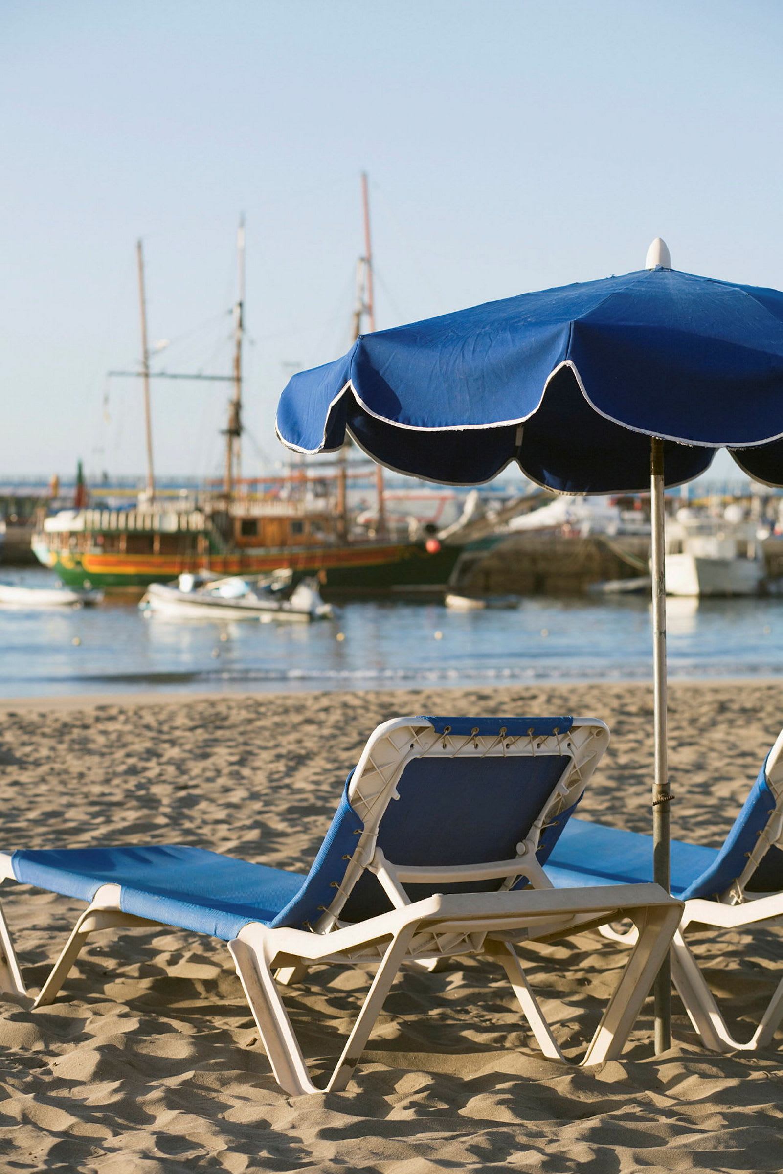 To solsenger med blå puter og en blå parasoll på en sandstrand med seilbåter i bakgrunnen. (AI-generert tekst)