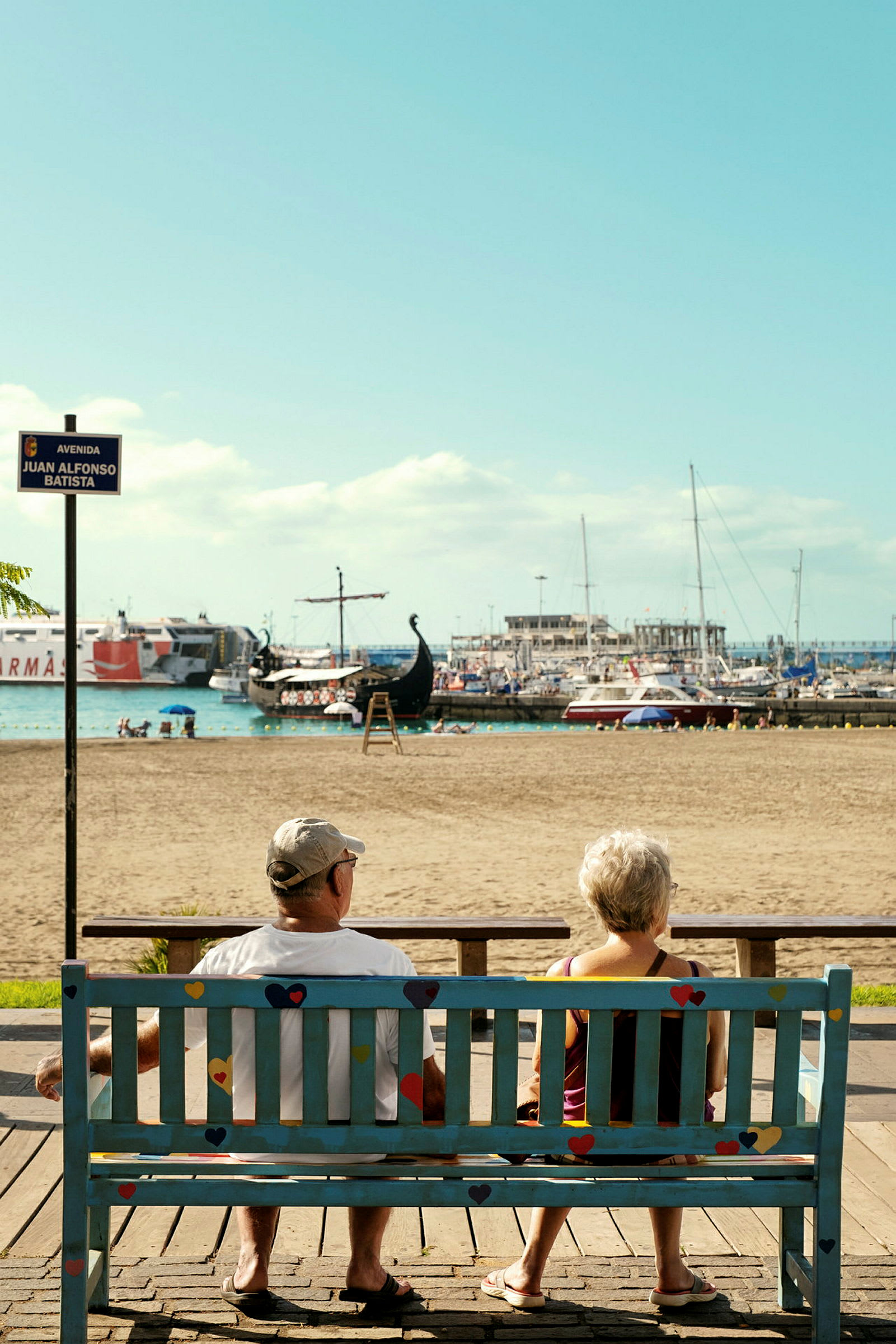 To personer sitter på en benk med utsikt over stranden og havnen. (AI-generert tekst)