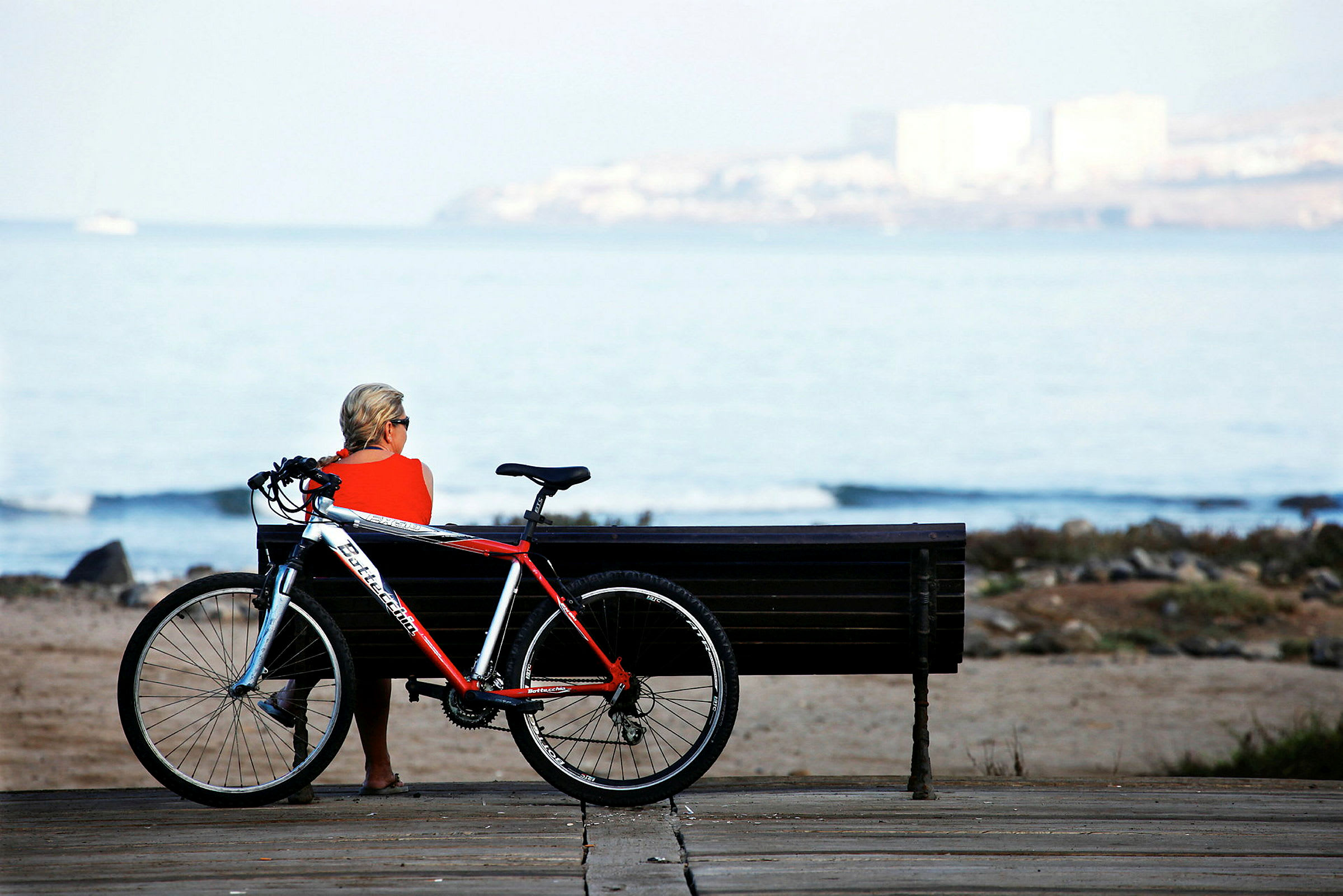 En person sitter på en benk ved stranden med en sykkel ved siden. (AI-generert tekst)