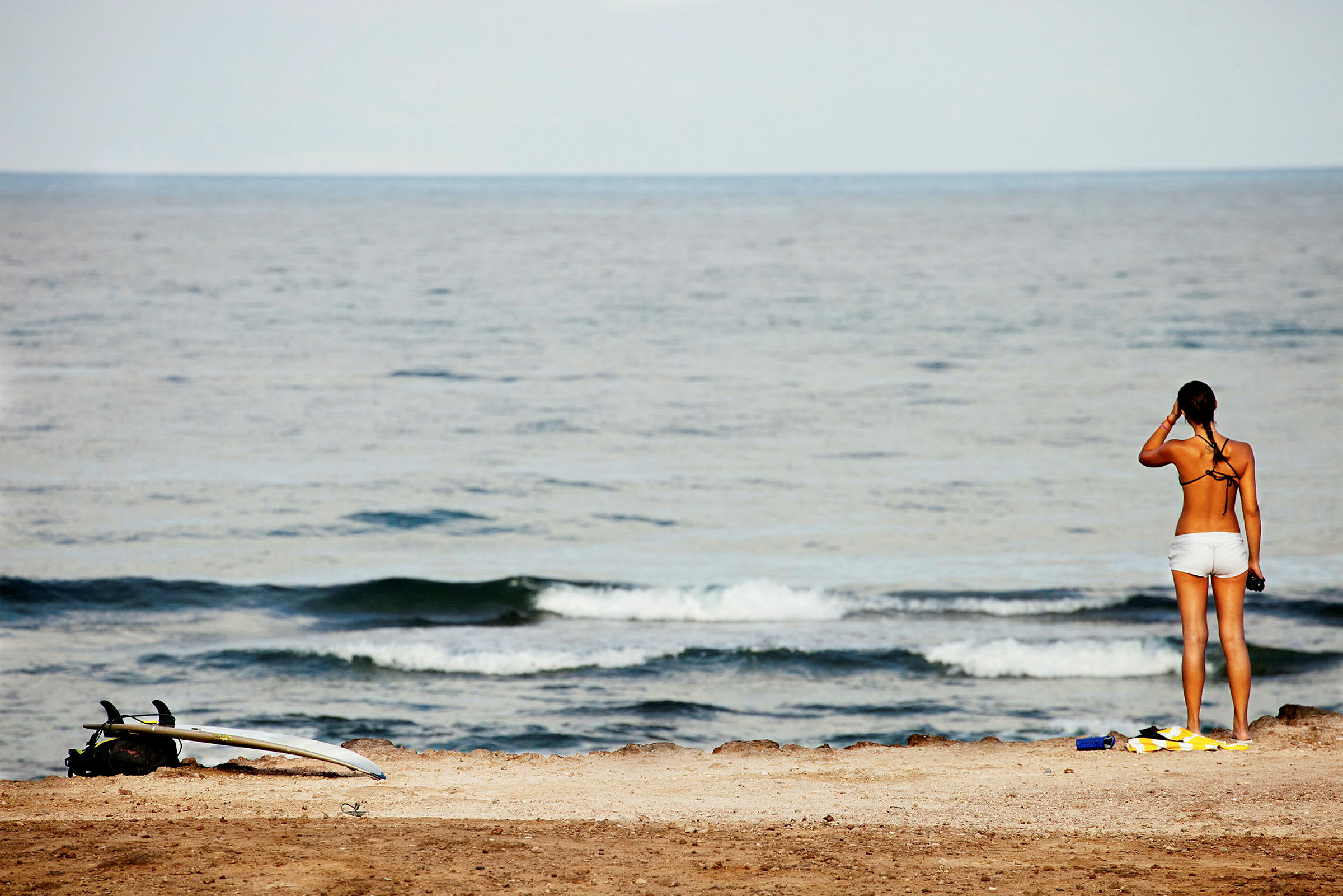 En kvinne står på en strand og ser utover havet nær et surfebrett. (AI-generert tekst)