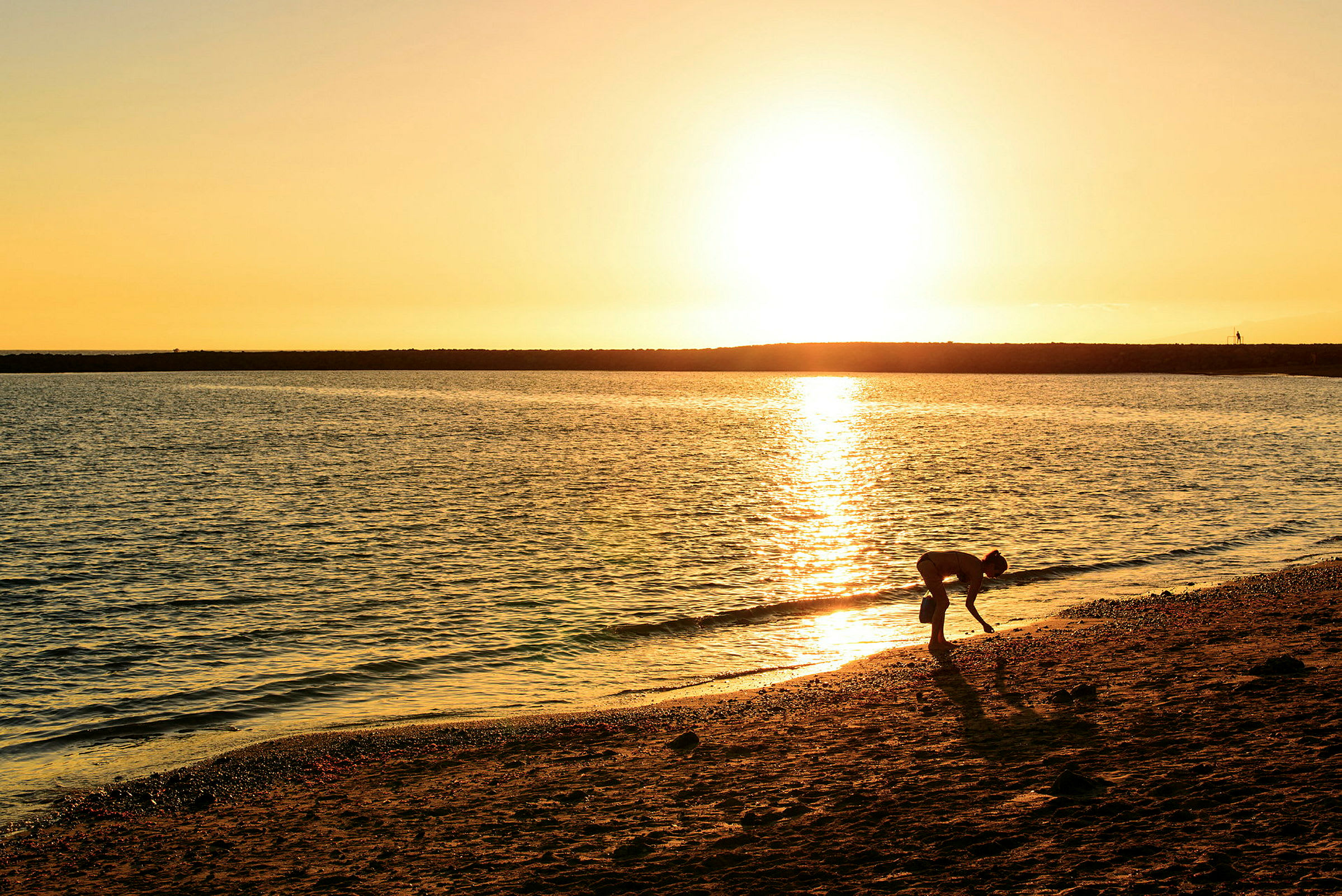En person plukker noe på stranden under solnedgangen. (AI-generert tekst)